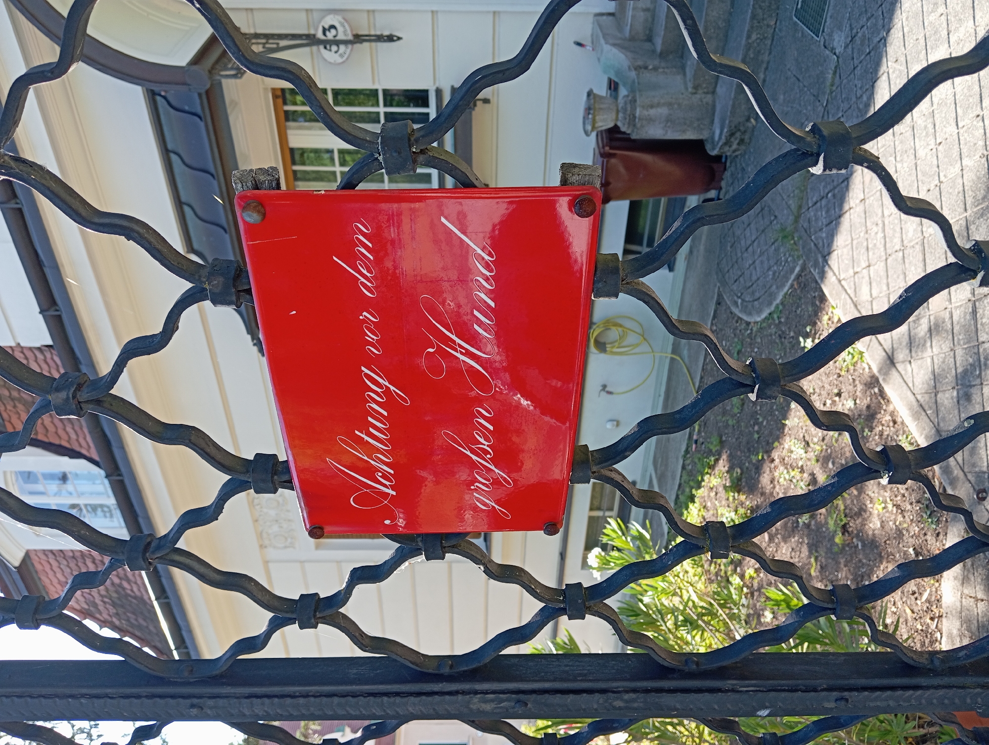 A red rectangular sign with white cursive text is affixed to a black wrought iron fence. The sign reads "Achtung vor dem großen Hund". The fence is in front of a cream-colored building with a red-tiled roof. A window with white shutters is visible on the building. The ground in front of the fence is paved with grey tiles and there is some greenery. The sign is attached to the fence with two rivets. The scene appears to be in a residential area. The building has a house number 53 visible on its facade.