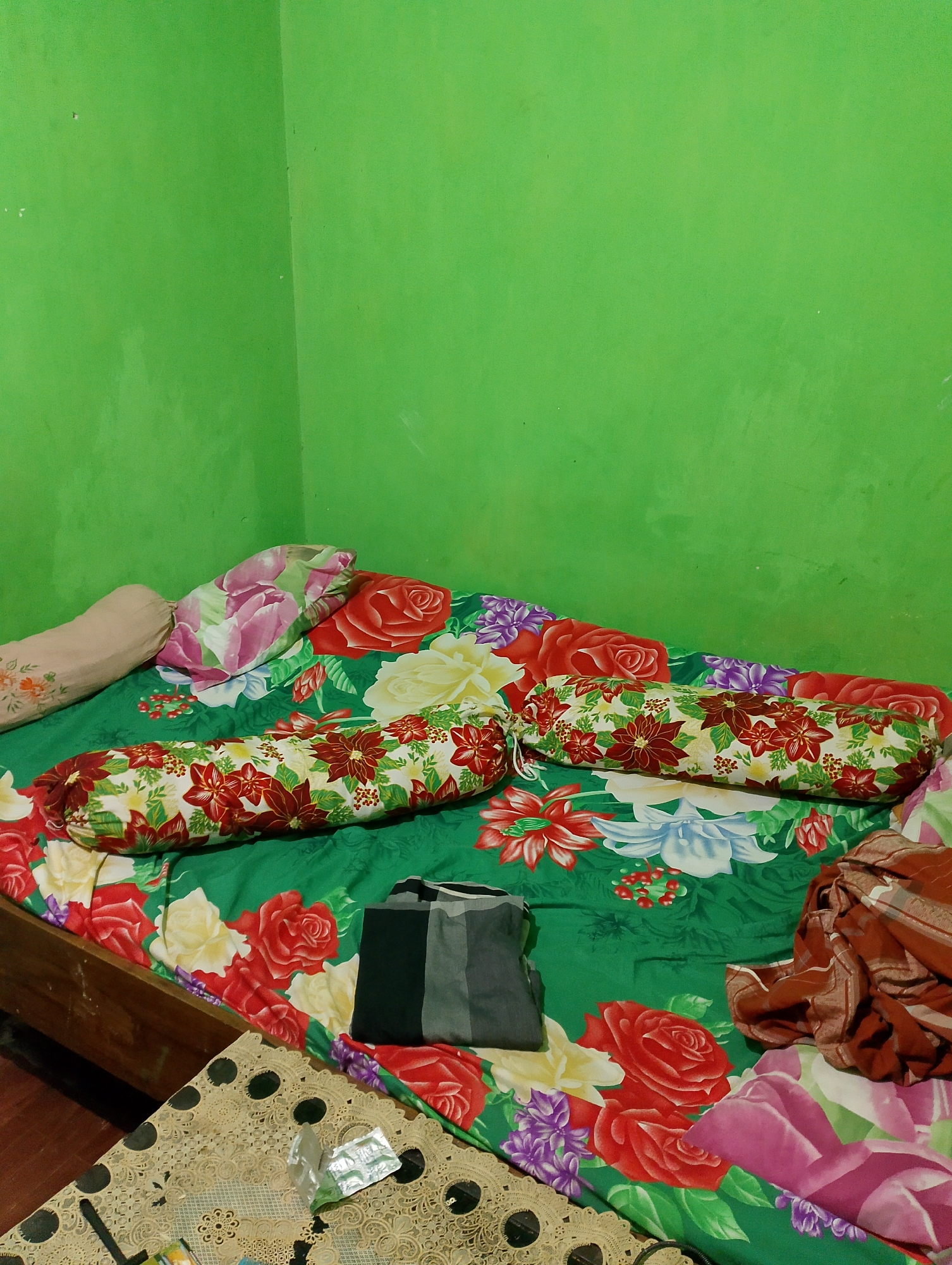 The image depicts a bed with a vibrant floral-patterned comforter and several pillows in a room with lime green walls. Two bolster pillows with a red and white floral pattern are positioned horizontally on the bed. A regular pillow with a pink and green floral design is tucked behind them. To the left, a tan bolster pillow with a subtle floral print is visible. A dark grey and black folded garment lies on the right side of the bed, near a pile of brown and white plaid fabric. In the foreground, a table covered with an ornate beige lace tablecloth is partially visible. A small, clear plastic wrapper with some contents is placed on the tablecloth. The wooden bed frame is visible on the left. The overall scene suggests a bedroom interior, likely in a domestic setting. The specific location in Rembang, Indonesia, is not discernible from the visual information provided in the image.
