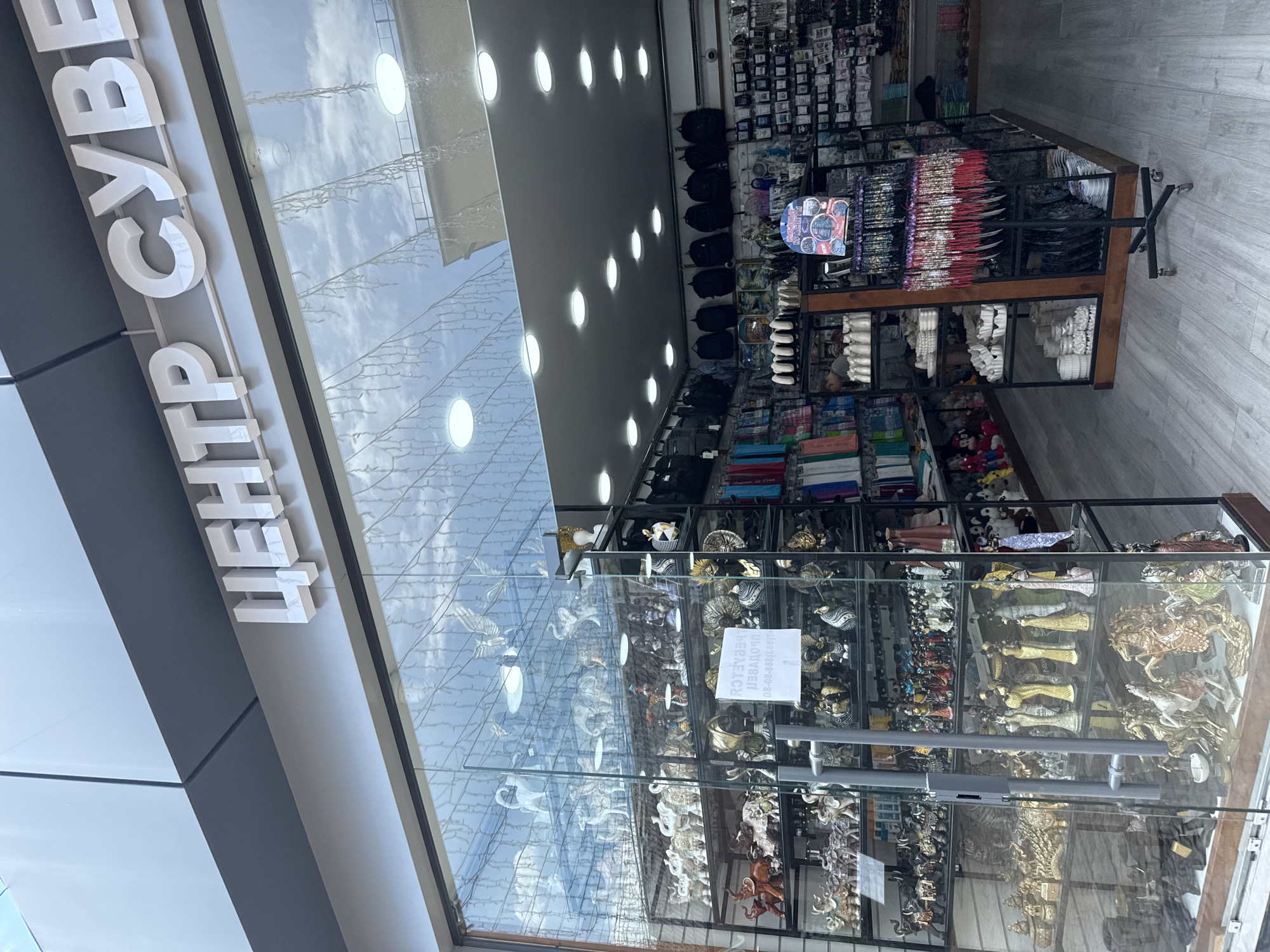 The image shows the exterior of a shop with a large glass window displaying various decorative items, primarily figurines of animals and people. Above the window, white, three-dimensional letters spell out "ЦЕНТР СУВЕ" in Russian, indicating "Center of Souvenirs." Inside the shop, shelves are filled with these decorative items, including numerous elephant figurines, statues of women, and horse sculptures. There are also bags and other merchandise visible on shelves and racks in the background. The shop appears to be located in a commercial area, possibly a street or an arcade, with a paved floor and overhead lighting. The reflection on the glass suggests an outdoor environment with a sky and buildings. The city of Sochi is mentioned in the prompt, and this type of shop is common in tourist destinations.