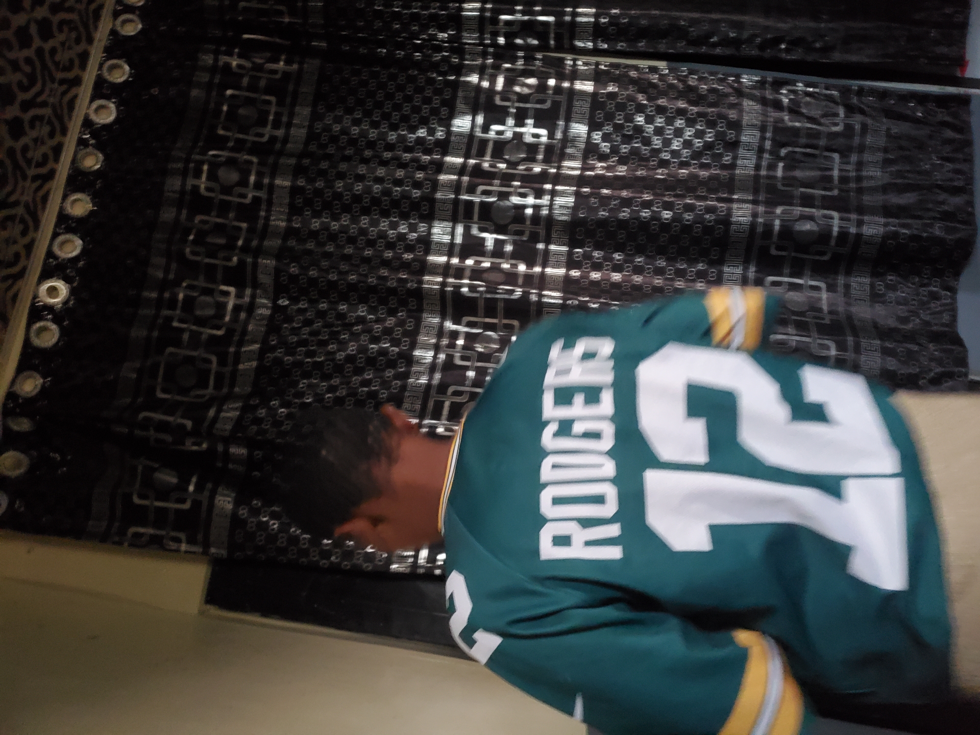 A person, seen from behind, wears a green football jersey with the name "RODGERS" and the number "12" in white on the back. The person's left arm is extended slightly, and their right arm is bent. The jersey has yellow and white stripes on the sleeves. The background consists of a black curtain with a repeating geometric pattern and lighter accents. The scene appears to be indoors, possibly in a residential setting, though the specific location in Winters, United States of America, cannot be determined from the image alone.