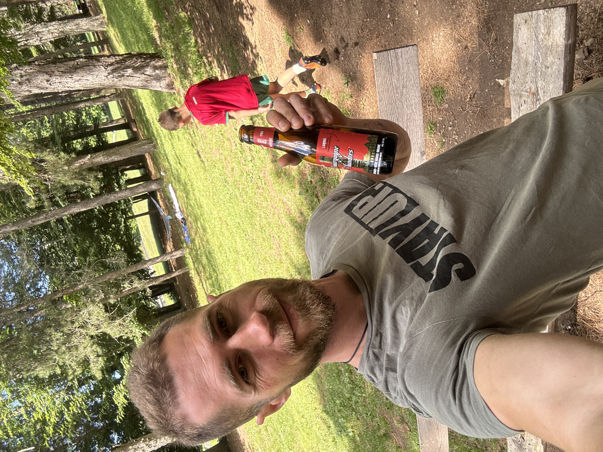 An adult male with light brown hair and a beard takes a selfie in an outdoor setting in Dolovi, Montenegro. He wears a dark olive green T-shirt with a black "STAY UP" graphic and a black cord necklace. In his right hand, he holds a brown glass bottle of "Bourgogne des Flandres" Lambic beer, with a red label indicating "ALC. 5% VOL". He smiles directly at the camera. In the midground, a young male child, wearing a red "PORTUGAL" shirt, green shorts, and colorful athletic shoes, walks away from the camera. The setting is a park or wooded area characterized by tall trees, green grass, and patches of reddish-brown dirt. A two-plank wooden bench is visible in the foreground, and further in the background, a light-colored blanket and a small ball are on the grass. The scene is brightly lit by daylight.