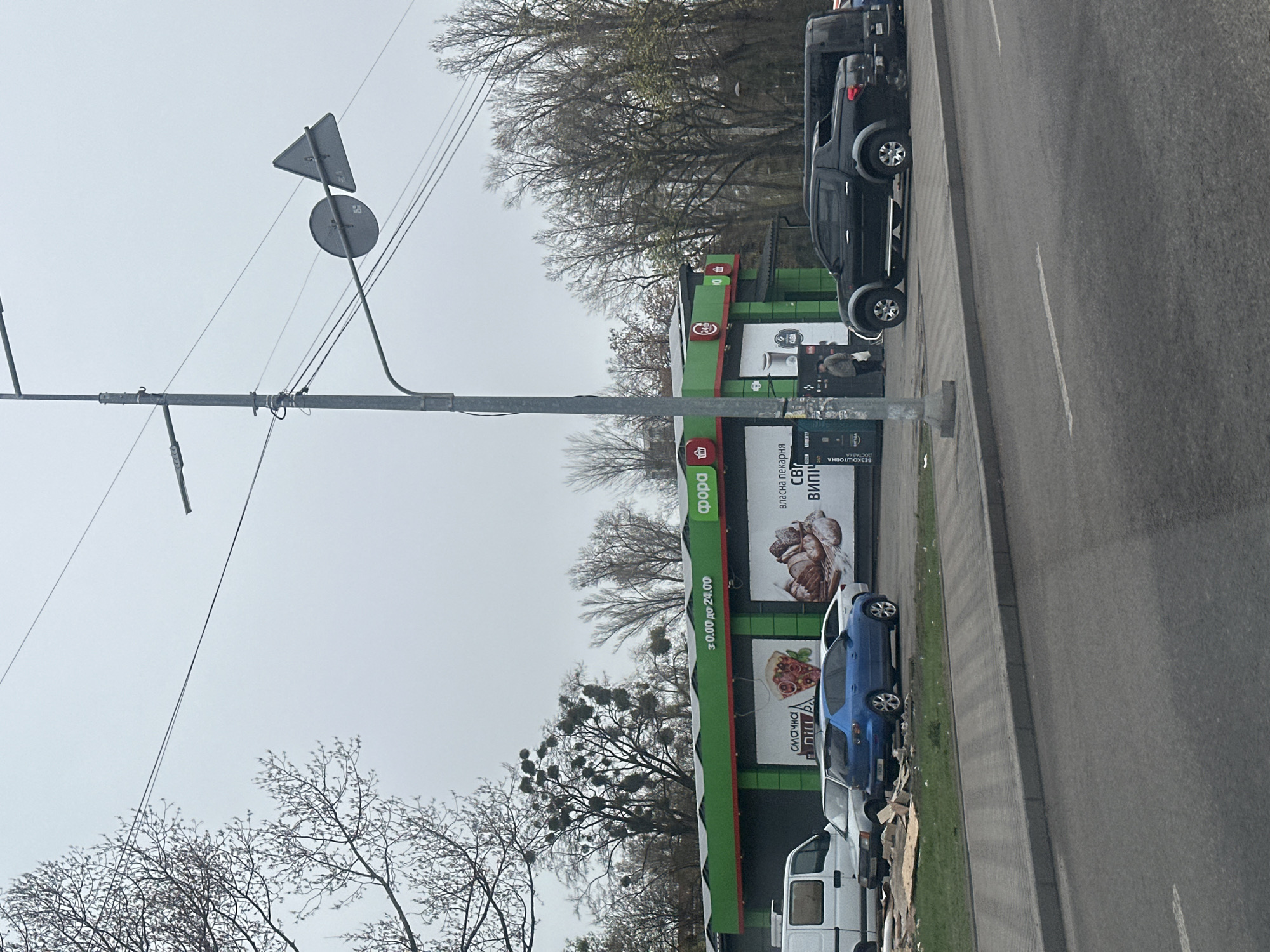 The image captures a street scene in Kyiv, Ukraine, showing a "Фора" (Fora) grocery store situated along a road with parked vehicles. The store's exterior is marked by green and red signage and advertisements for baked goods and pizza. Parked cars, including a dark-colored pickup truck, a blue sedan, and a white van are situated along the side of the road beside the building.
Utility poles with overhead wires are also visible. The sky is overcast, and bare trees suggest it is either late autumn or early spring. The scene appears to be an ordinary street with commercial establishments and parked vehicles.