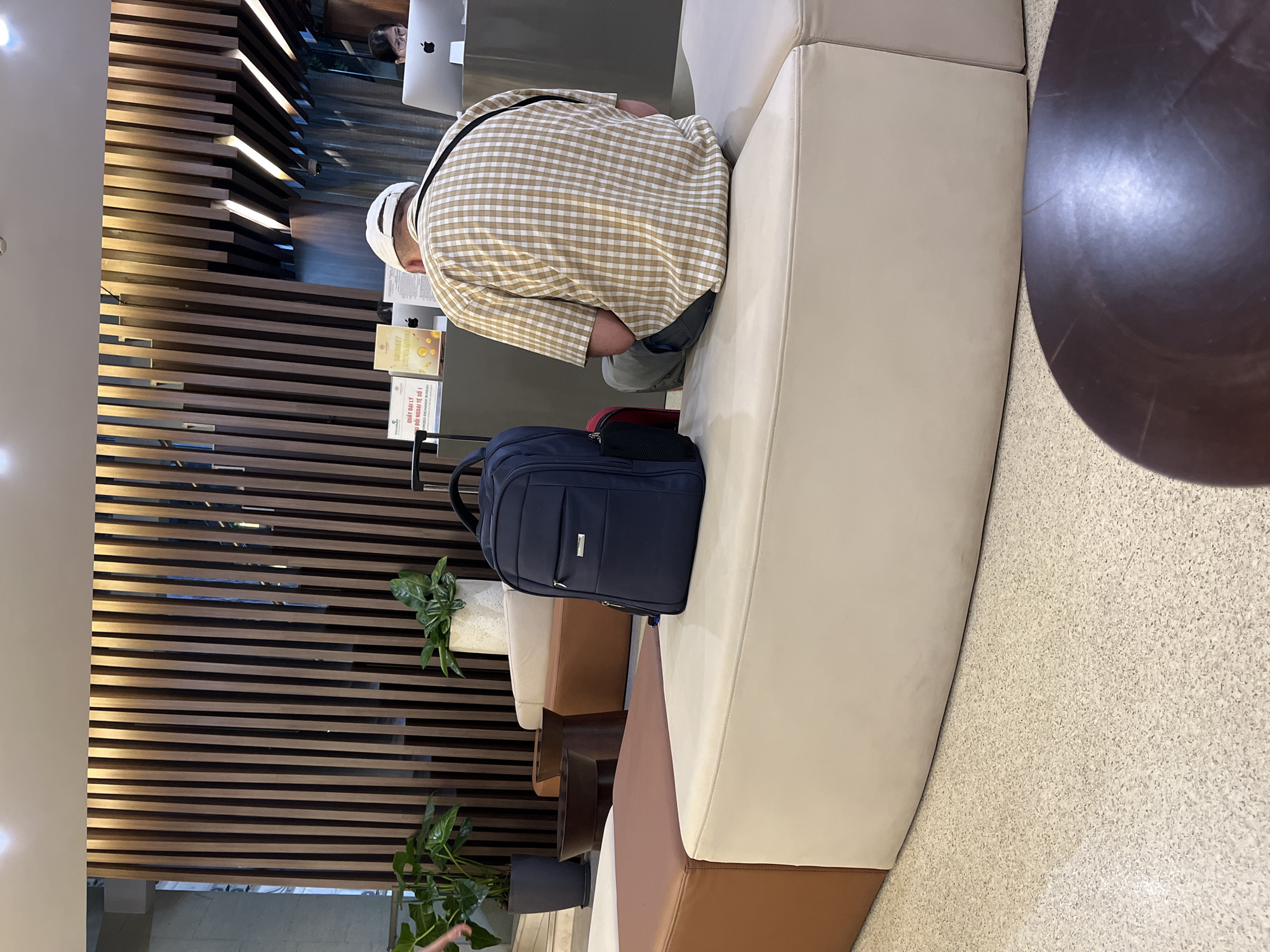 The image shows a man sitting on a curved, layered sofa with a dark blue backpack next to him. The setting appears to be a public space, possibly a waiting area or lobby, given the presence of plants, a textured wall, and what seems like a reception desk or computer terminal in the background. The man is wearing a checkered shirt and a white hat. The location is in Đà Nẵng, Vietnam, as indicated by text on a nearby sign. A round table is positioned next to the sofa. The floor has a speckled pattern.