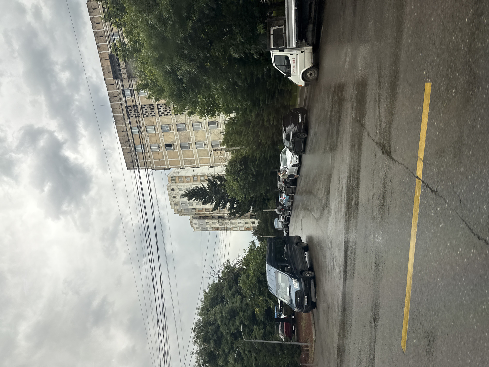 An overcast, gray sky looms above a street scene in an urban environment. Tall, beige apartment buildings with numerous windows and some visible facade damage stand on the right side of the image, partially obscured by lush green trees. A row of parked cars, including a dark blue van in the foreground, a white sedan, and a black car, are lined up on the wet asphalt road. A white flatbed truck is also parked on the right side of the road. The road surface appears wet, suggesting recent rain, and is marked with a yellow line. Utility wires are visible strung across the sky. The overall atmosphere is somber and subdued due to the cloudy weather.