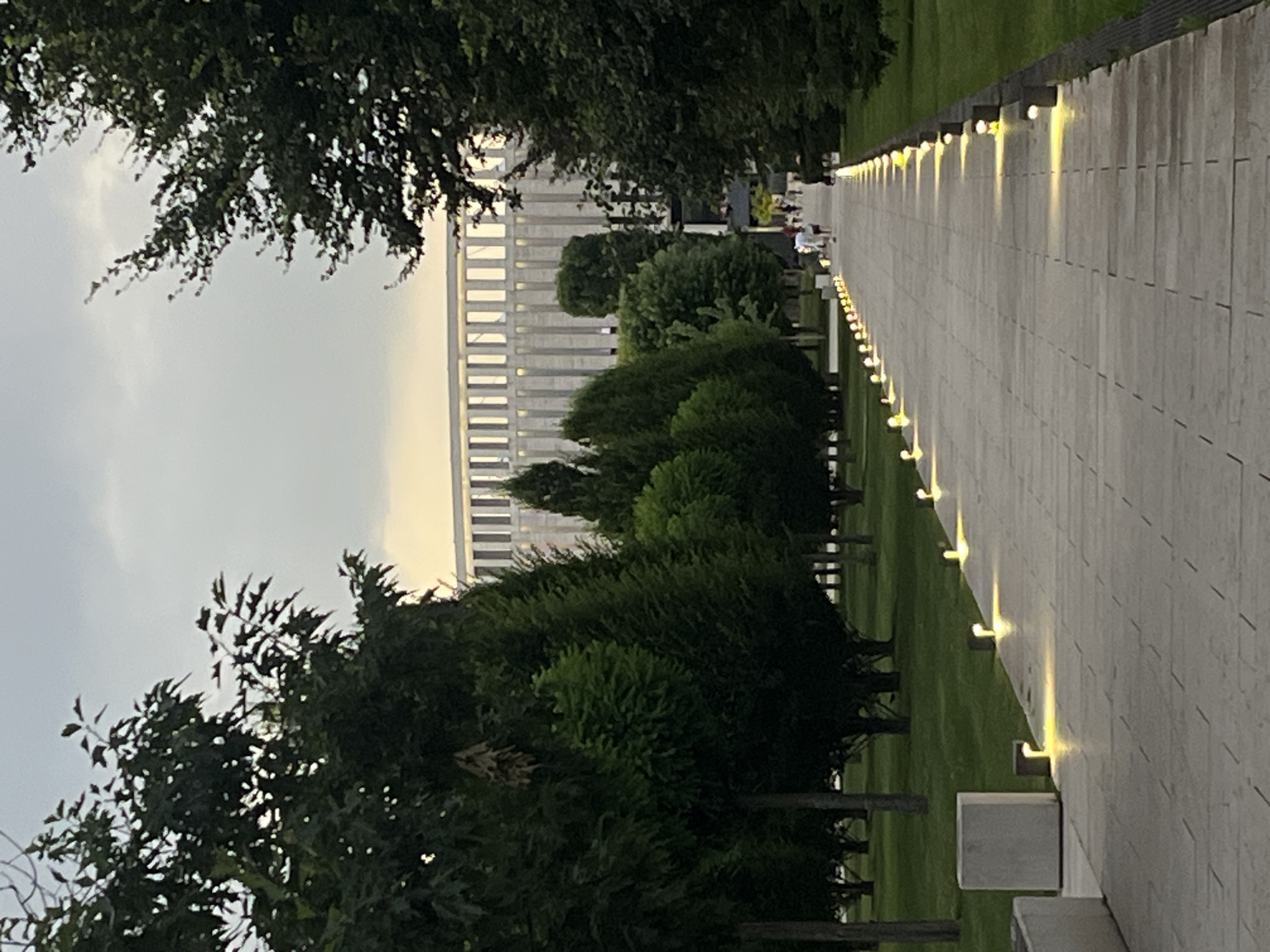 The image depicts a pathway lined with lights and trees, leading towards a large, modern building. The pathway is paved with light-colored rectangular stones, and small, upward-facing lights are intermittently placed along its edge, casting a warm glow. To the left of the pathway, there is a well-maintained lawn dotted with trees of various sizes and shapes. In the distance, a multi-story building with a distinct, repetitive architectural style is visible. The sky is overcast, suggesting either early morning or late afternoon. There are a few people walking along the pathway in the distance, and some people are seated at tables in an outdoor area near the building. The overall scene suggests a public park or a memorial site. There is no visible text. The location context provided is Krasnodar, Russia.