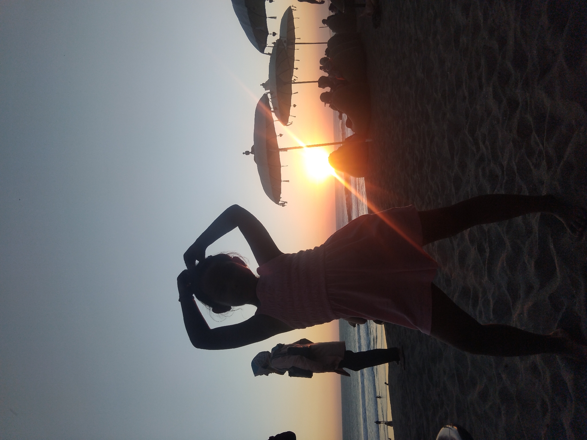 Viewed horizontally, the image captures a serene sunset scene on a beach, likely in North Kuta, Indonesia. The primary subject is a young girl in a pink dress, rendered in silhouette against the vibrant sky. She stands with her hands raised and clasped behind her head, striking a pose or stretching.
Behind the girl, several large, traditional beach umbrellas are silhouetted against the bright evening light. The sun, a radiant orange orb, is prominently positioned just above the horizon, situated between two of these umbrellas and casting dramatic lens flares across the scene. The sky transitions from brilliant orange and yellow near the horizon to a soft blue-grey higher up.
Further in the background, the ocean meets the shore, with gentle waves visible. Several other silhouetted figures can be seen along the waterline and on the sandy beach, observing the sunset or enjoying the atmosphere. The sand in the foreground displays subtle undulations. The image portrays a tranquil moment at dusk, emphasizing the beauty of the natural light and the relaxed ambiance of the beach. No text is visible in the media file.