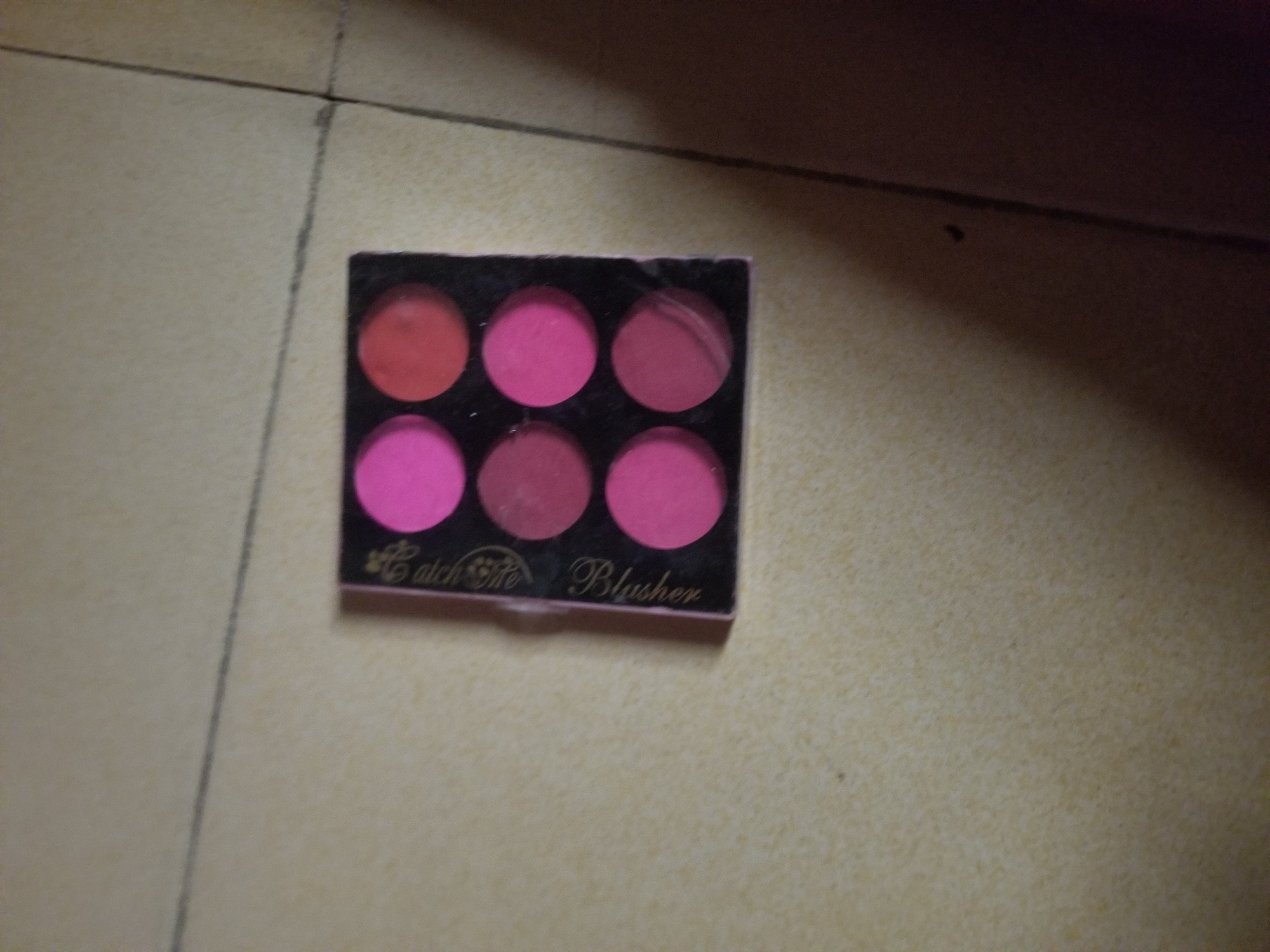 The image shows a palette of six round blush pans arranged in two rows of three. The blushes are various shades of pink and reddish-pink. The palette itself is black and rectangular. The text "Catch Me Blusher" is visible on the front of the palette. The palette is placed on a textured, light-colored surface that appears to be tiled flooring, with a thin dark line running diagonally across it. The lighting in the image is dim, with shadows suggesting an indoor setting. The overall scene is a close-up shot of the makeup product.
