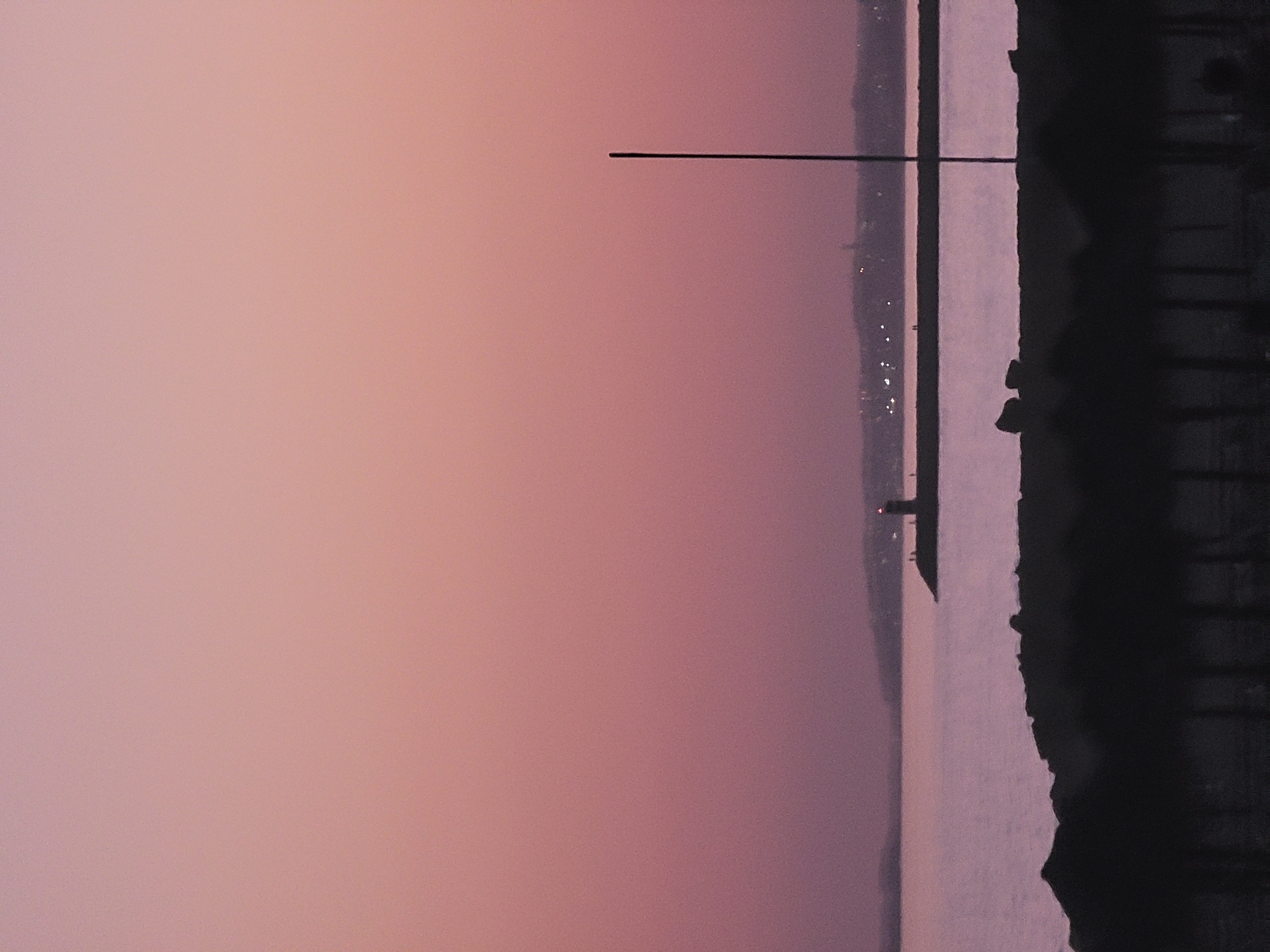 The image depicts a serene coastal scene at dusk in Quarteira, Portugal. The sky is a soft gradient of pink and purple hues, suggesting the last light of day. In the foreground, silhouetted against the twilight, are the shapes of beach umbrellas and rocky formations. The mid-ground features a calm body of water. Further out, a pier juts into the sea, and on the end of this pier stands a lighthouse with a red light illuminated at its top. A distant, faint line of city lights is visible along the horizon, adding a touch of urban presence to the natural landscape. A single, thin pole stands vertically in the right mid-ground, silhouetted against the sky.
