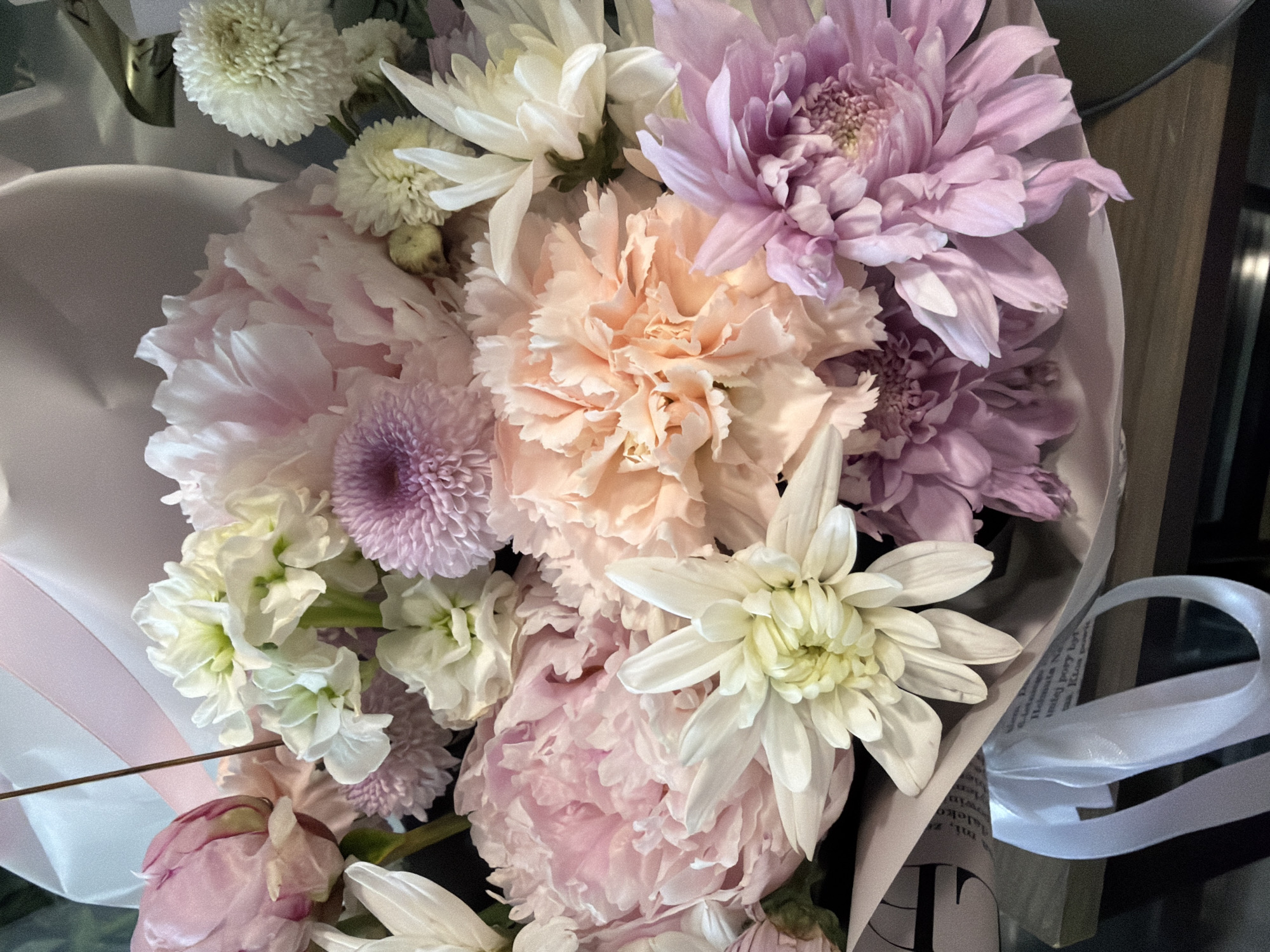 A close-up overhead shot shows a bouquet of flowers with soft pink, white, and peachy-orange tones. The flowers are varied in type and include what appear to be peonies, carnations, and chrysanthemums. The bouquet is wrapped in light pink and white paper, and a white ribbon is tied around the lower part. The flowers are arranged densely and fill the frame. The background is dark and indistinct, suggesting it might be a dimly lit indoor setting. There are no people or discernible location details.