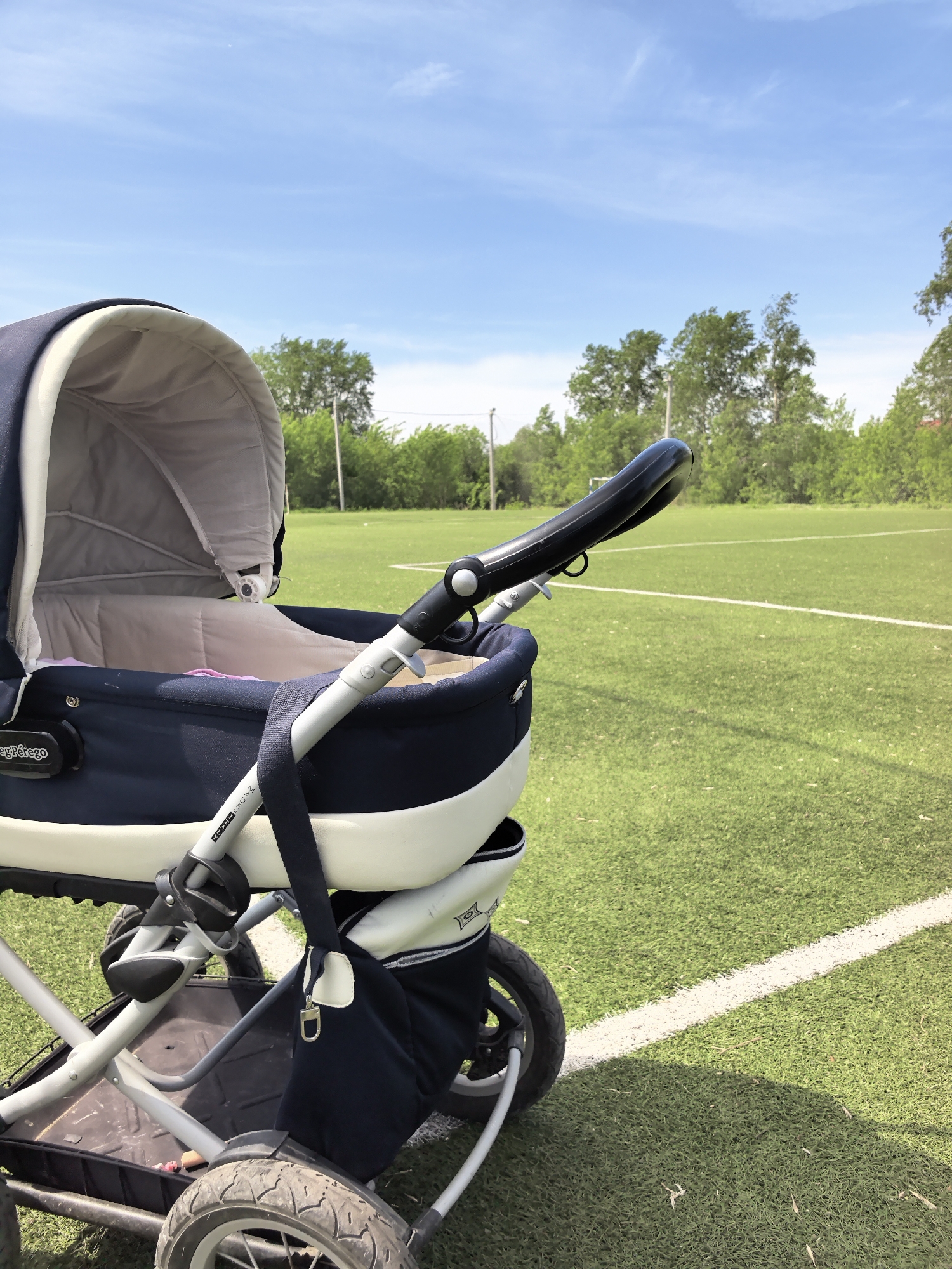 The image depicts a baby stroller positioned on the artificial turf of a sports field. The stroller is a Peg Perego model, navy blue and cream in color. The stroller faces right in the frame, with the handle extending toward the background. The field is green with visible white lines, indicating it's likely used for soccer or similar sports. Beyond the field, there is a line of trees. The sky is blue with light clouds. The location is Фролы, Russia. No people are visible in the image.