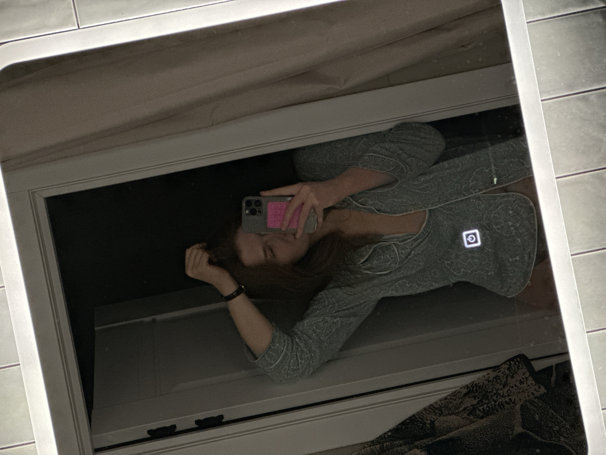 A woman is visible in the reflection of a lit mirror. She is holding a smartphone and taking a selfie. The background suggests a bathroom, with tiled walls visible around the mirror. The woman is wearing patterned clothing. The location cannot be determined from the image.
