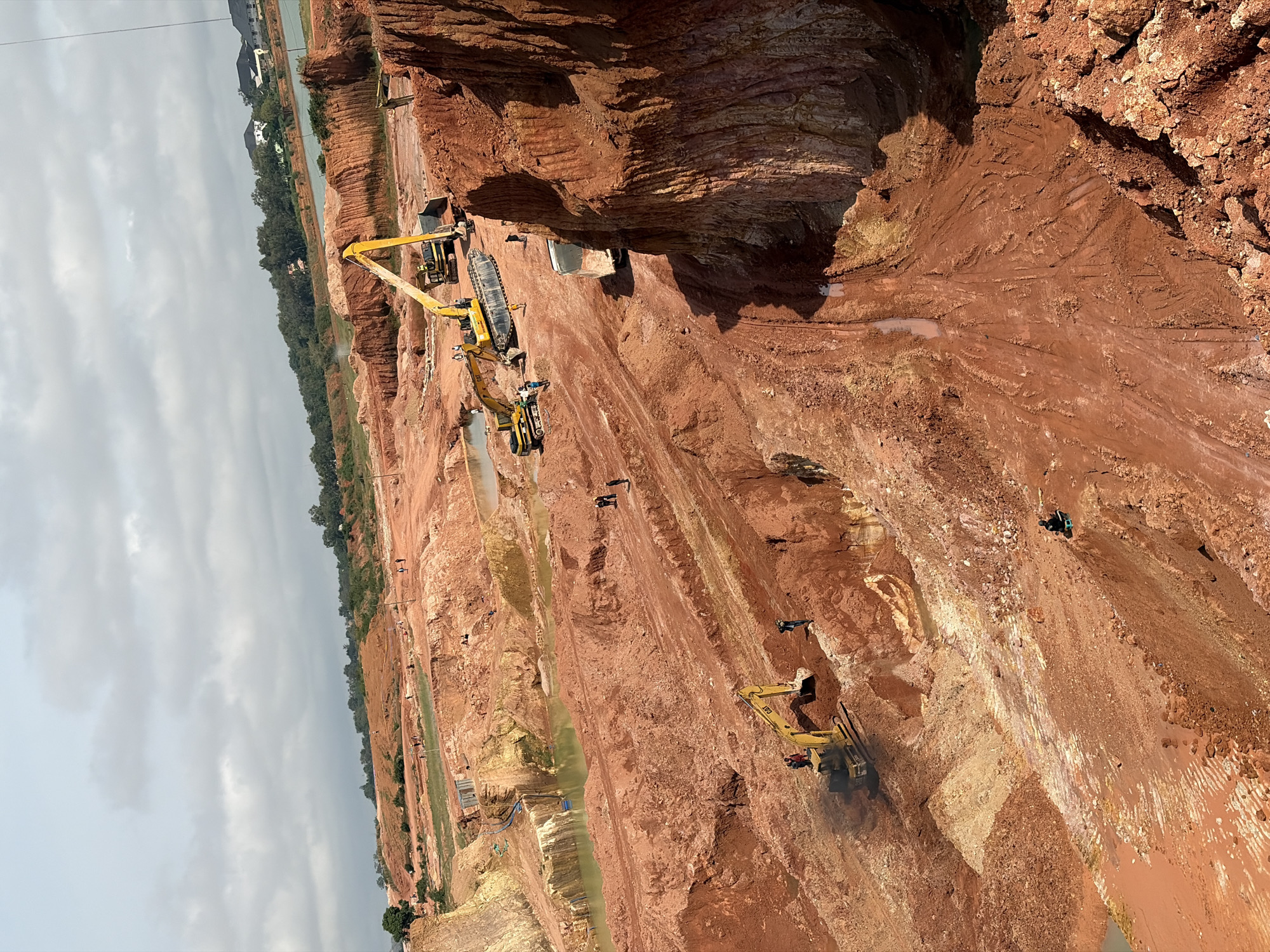 The image depicts an expansive open-pit mine characterized by large excavations and piles of reddish-brown earth. The terrain is rugged and uneven, with sheer drops and layered strata of soil and rock. Several heavy construction vehicles, including yellow excavators and a large dump truck, are actively engaged in excavation and transportation. Numerous individuals, dressed in casual attire or construction gear, are scattered throughout the site, some operating machinery, others walking or standing near the equipment. The environment includes areas of standing water within the excavated pits, reflecting the sky. In the background, a more developed area with houses and trees borders a body of water, suggesting the proximity of a settlement to the mining operation. The sky above is overcast with scattered clouds. The scene is indicative of ongoing mining or earth-moving activities within a substantial geological formation, likely in an area undergoing significant land modification. This scene is consistent with active quarrying operations, possibly for raw materials such as clay or sand, in the region of Kwang, Nigeria.