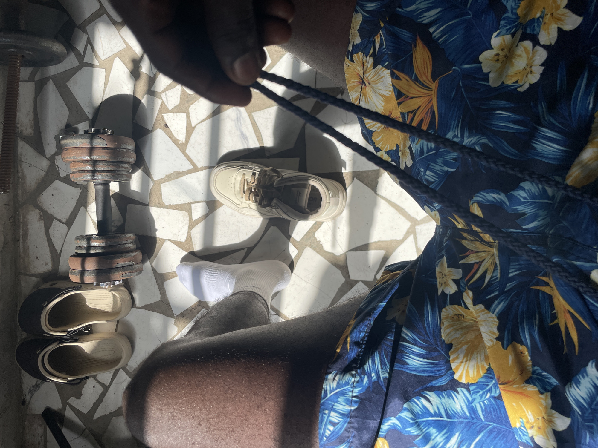 The image, captured from a first-person perspective looking down, shows a person's legs, one arm, and hand in a sunlit indoor or semi-outdoor setting in Badagry, Nigeria. The person is wearing dark blue shorts with a vibrant tropical pattern featuring yellow, light blue, and white floral and leaf motifs. One hand is visible, holding a dark, braided cord or drawstring, possibly from the shorts. One foot is visible, wearing a white sock.
On the irregular white-tiled floor, several objects are arranged. To the left, an old, adjustable dumbbell with multiple rusty, dark weights is visible. Next to the dumbbell, a pair of black and beige clogs or sandals are stacked. Towards the center of the frame, a single beige sneaker is placed on the tiles.
Strong, direct sunlight casts sharp, elongated shadows across the tiled floor, creating distinct light and dark patterns. The irregular, light-colored tiles have darker grout lines, giving the floor a rustic or aged appearance. The dumbbell weights show signs of wear and rust. There is no discernible text visible in the image.