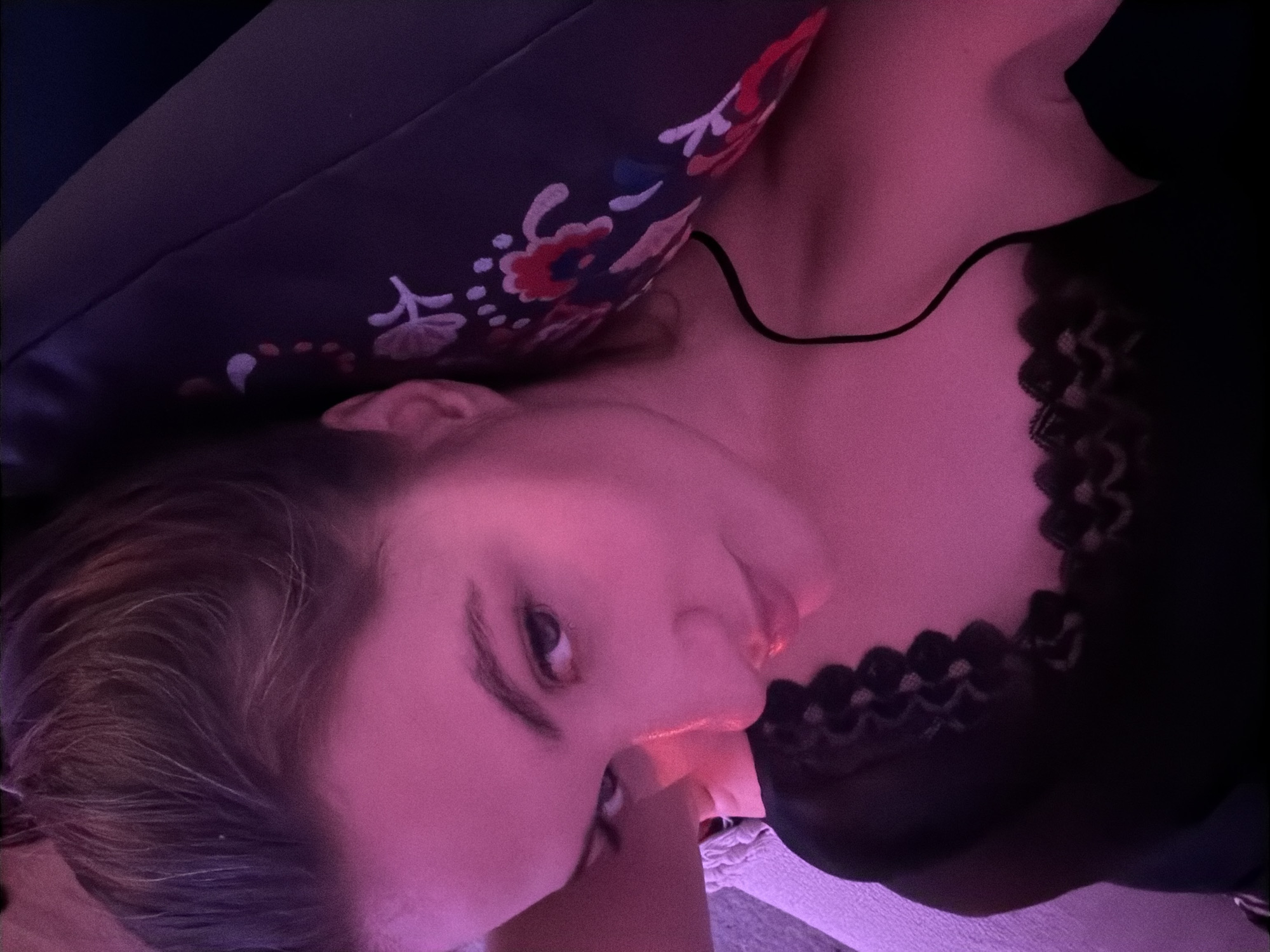 An indoor scene, presumably in Belgrade, Serbia, features a female subject from the chest up, positioned against a dark patterned cushion. The subject, a young adult with dark hair pulled back, gazes directly at the viewer. Her skin exhibits a strong magenta tint from ambient lighting. She wears a black garment with lace detailing along the neckline and a visible thin black shoulder strap. A dark cable rests across her right clavicle, extending off-frame. To the left, a light-colored, textured fabric surface is visible. The cushion, dark with white, red, and blue floral patterns, is situated behind her head and upper body. The immediate background is dark and undefined.