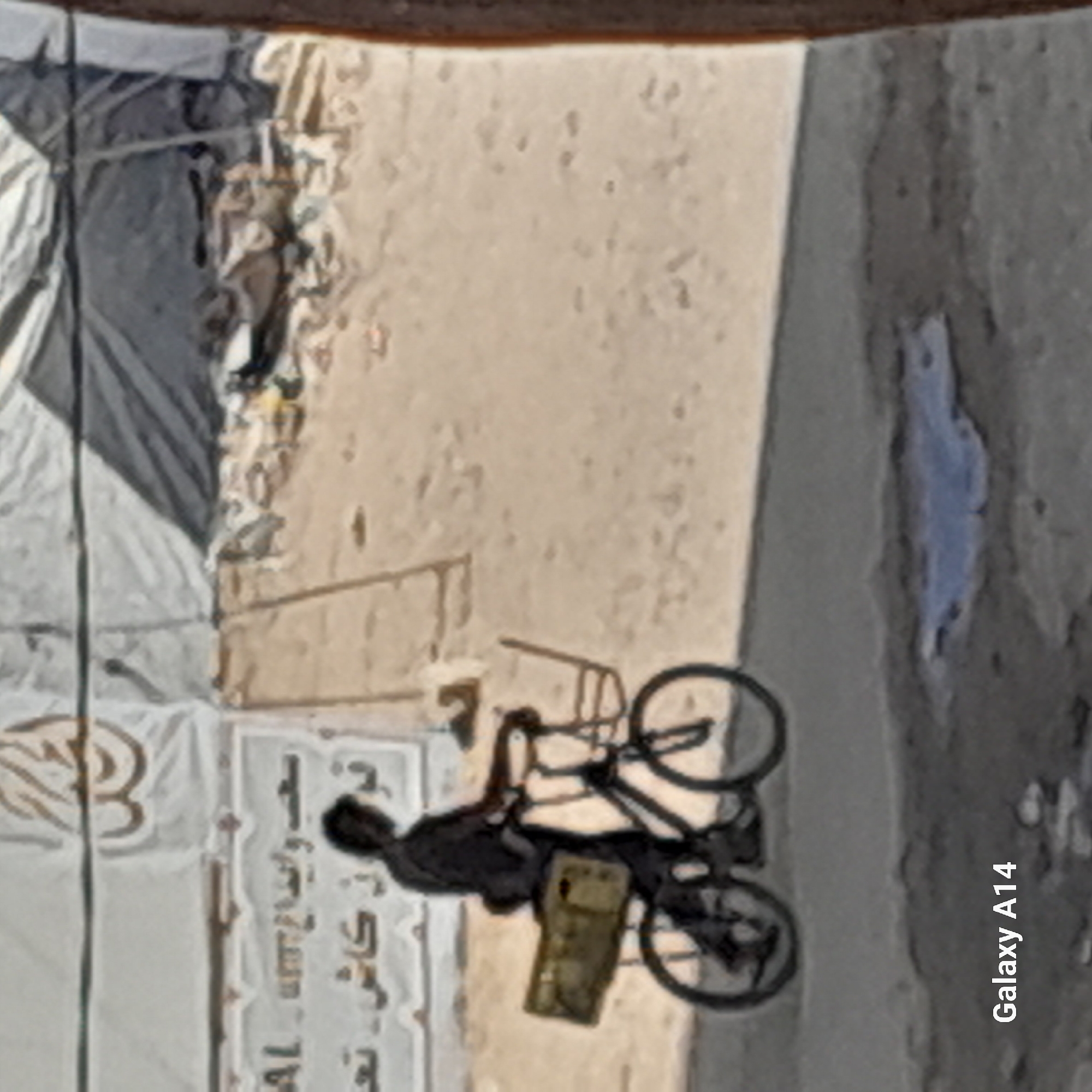In Khan Yunis, Palestinian Territories, a person is seen riding a bicycle on a dirt road. The person is wearing dark clothing and has a bag strapped to the back of the bicycle. In the background, there are tents and a banner with Arabic writing. The ground is dry and dusty, with some debris scattered around. The overall scene suggests a rural or temporary settlement environment.