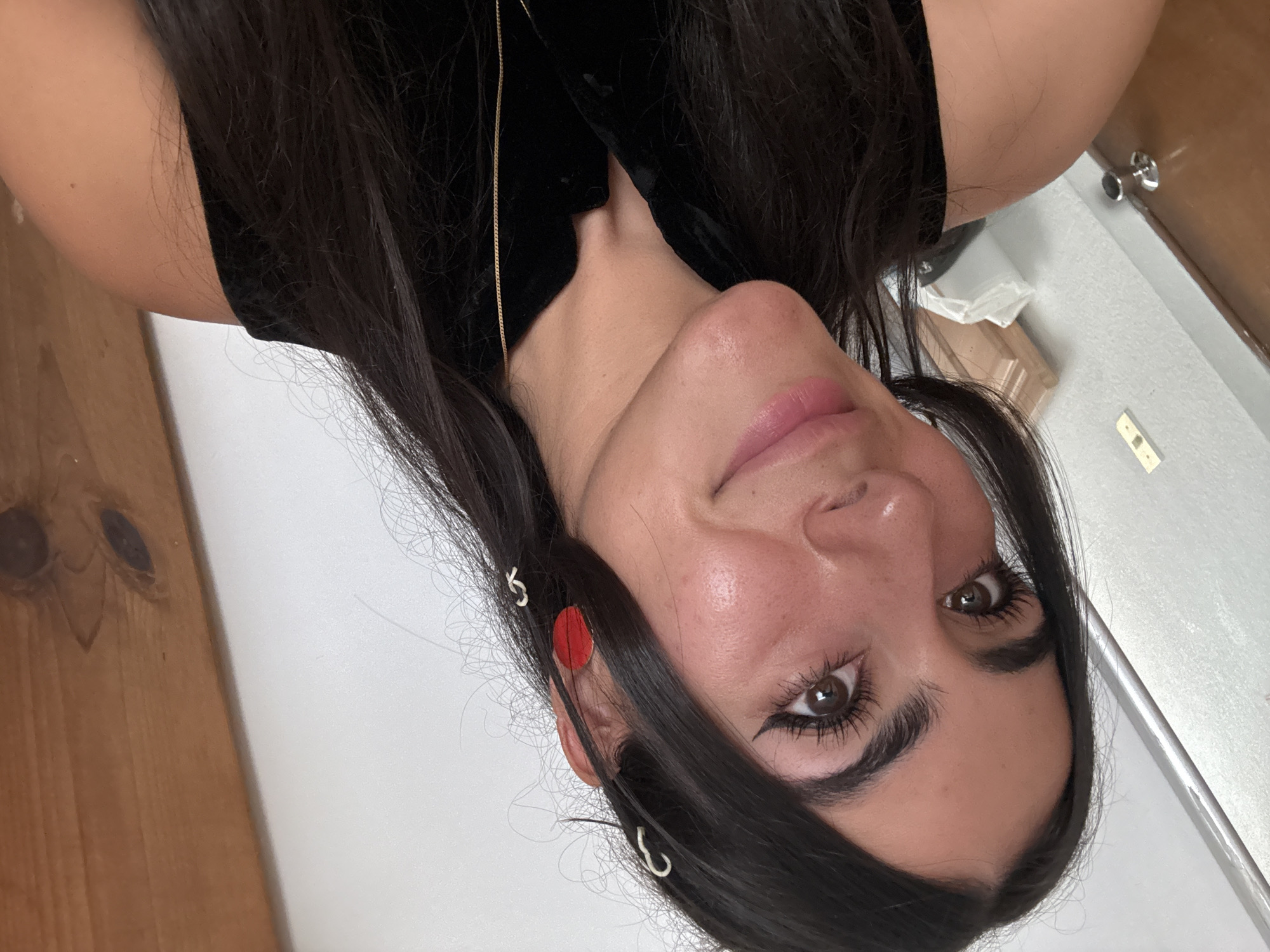 A close-up, correctly oriented portrait features a young woman with dark hair, looking directly at the camera with a subtle smile. Her head is tilted gently to her left, showcasing her right side more prominently. She has dark, expressive eyes enhanced with winged eyeliner and mascara, and her lips are adorned with light pink lipstick, giving her a natural and fresh appearance. Her long, dark brown hair frames her face, with several strands styled behind her right ear using small white spiral clips and a distinctive red circular clip. A delicate gold chain necklace is visible against her skin, peeking from beneath her black velvet sleeveless top. The background on the left consists of a light brown wooden surface, possibly paneling or a door. The rest of the background is a plain white wall, with a silver doorknob or handle and a light switch visible on the right side. The indoor setting appears casual and domestic. No visible text is present in the image.