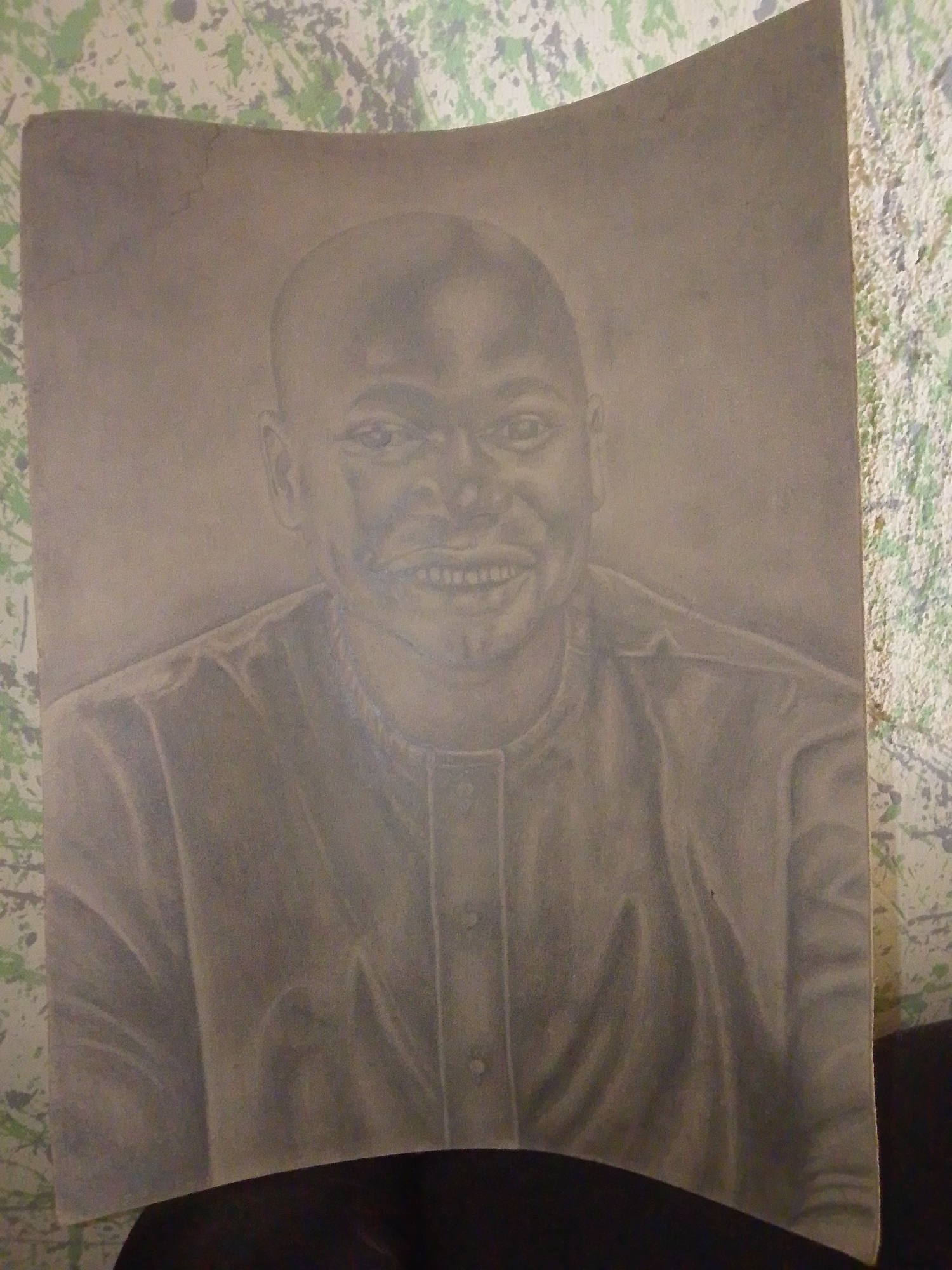 This is a photographic image of a charcoal drawing of a man's portrait. The drawing is on a light-colored paper that is slightly warped, with some faint lines and smudges visible. The man in the drawing has a shaved head, prominent ears, and is smiling broadly, revealing his teeth. He is wearing a collared shirt with buttons. The background of the drawing is a plain, light gray. The drawing is placed against a wall with a speckled green and white pattern. The image appears to be taken in an indoor setting, possibly a home or studio, in Jalingo, Nigeria. The lighting suggests it is daytime, but the exact source of light is not evident. There are no other people or objects clearly visible in the frame beyond the drawing and the wall it is leaning against. The overall presentation is informal.