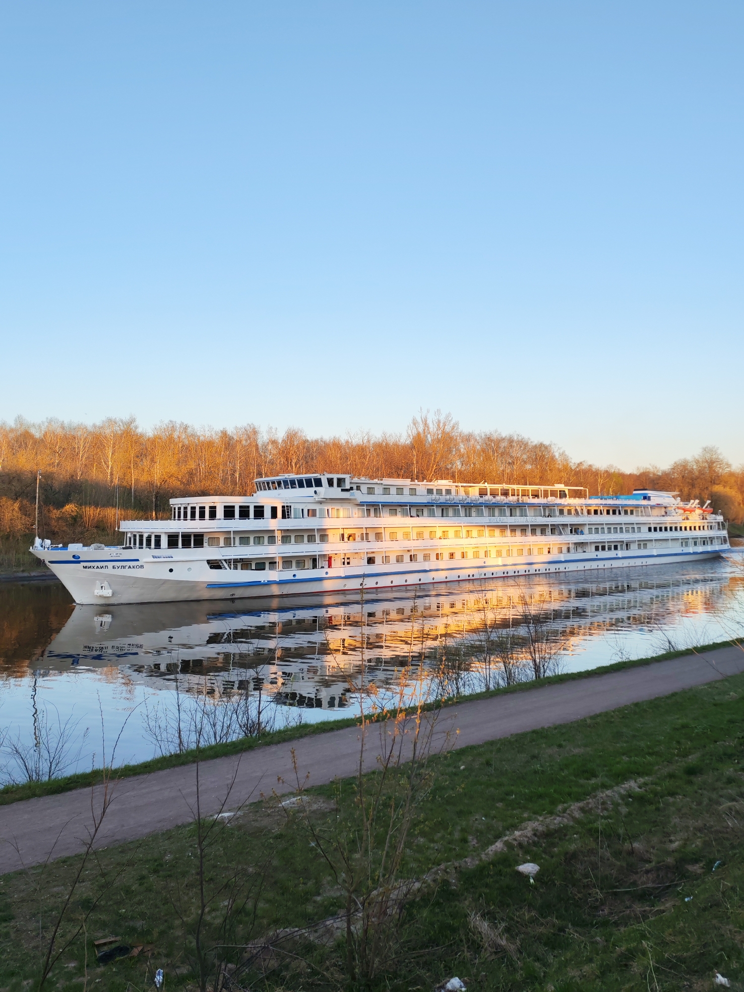 A long white passenger ship, named "Михаил Булгаков" (Mikhail Bulgakov), is traveling on a canal in Yakhroma, Russia. The ship has multiple decks and windows. The reflections of the ship and surrounding trees are visible in the water. A paved path runs alongside the canal. Grass and sparse vegetation cover the foreground. Bare trees are in the background. The sky is clear and light blue. The scene is illuminated by soft, golden light, suggesting either sunrise or sunset.