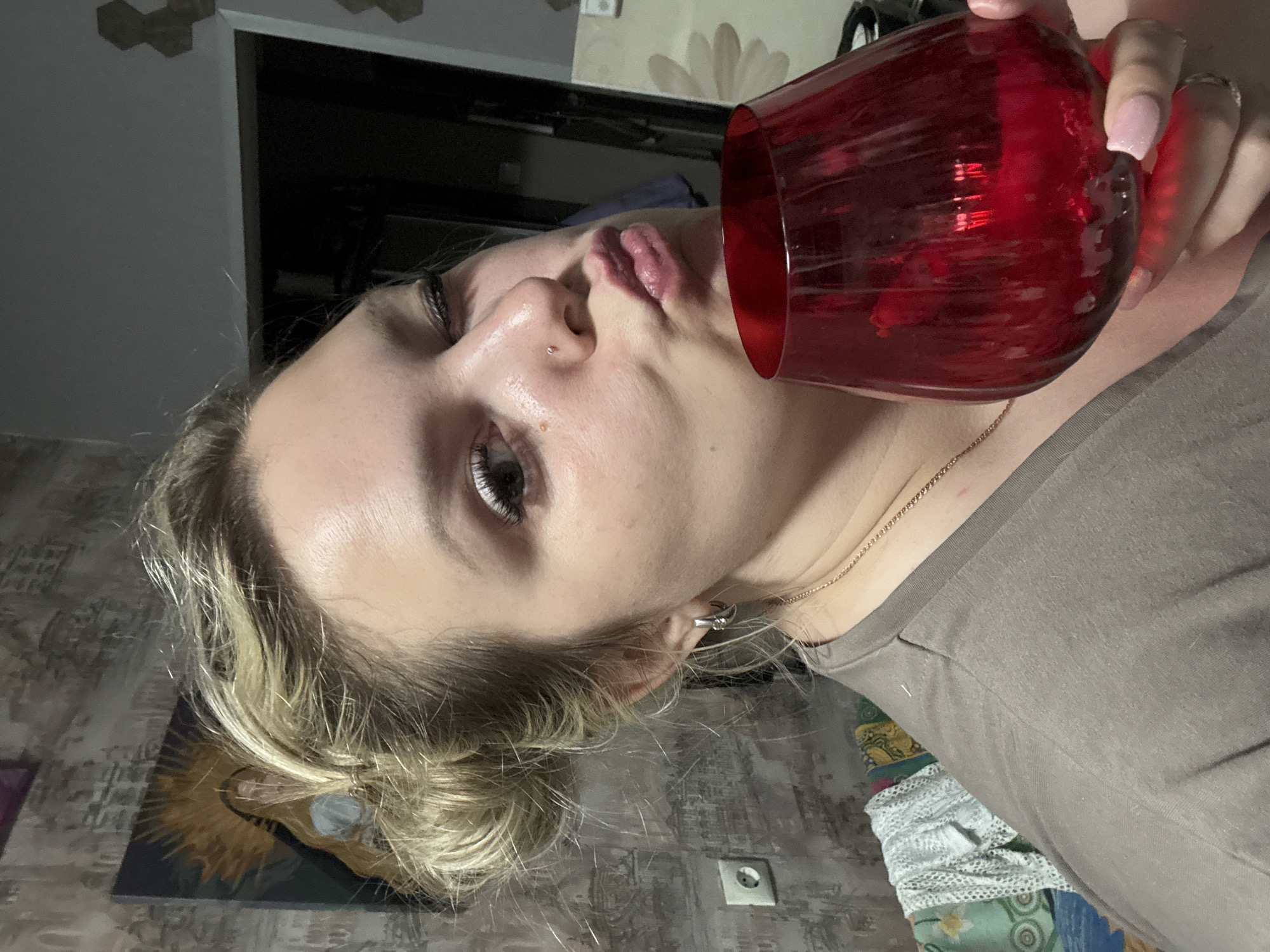A woman is drinking from a red glass. She has fair skin, blonde hair pulled back, and a nose piercing. The background includes a door, a picture frame, and an electrical outlet. The location is identified as Марковский, Russia.