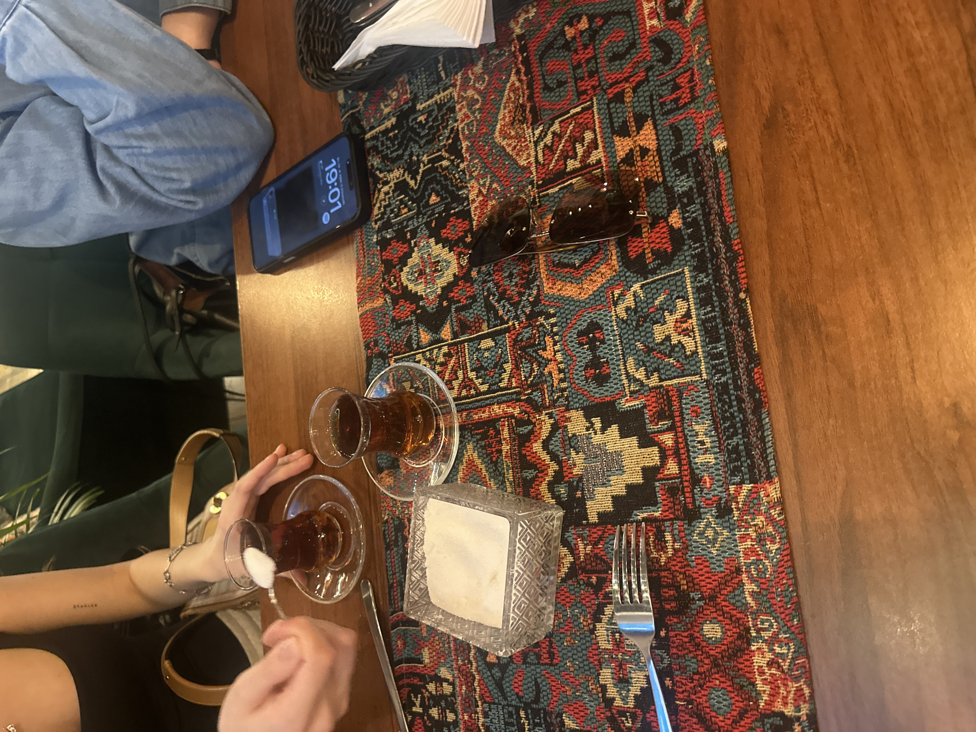 On a wooden table in an unknown location, two people are seated, one stirring sugar into a tulip glass of tea with a spoon. Two tulip glasses of tea sit on the table with saucers, next to a clear container holding white sugar cubes, and a metal fork. A colorful rug or runner covers part of the table. Sunglasses and a black smartphone are on the table, next to a basket containing paper napkins. One of the people is wearing blue jeans and the other is wearing a black shirt and has a tattoo on their forearm.