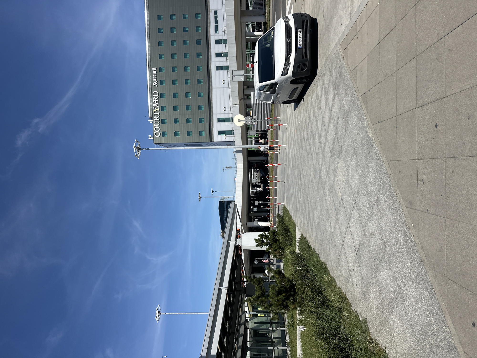This is a horizontal, eye-level, outdoor photograph taken on a sunny day in Warsaw, Poland. The image shows a modern building complex with a "Courtyard Marriott" sign. In the foreground, a white van with a license plate WPR 3114X is parked. The ground is paved with large grey tiles. A series of red and white striped bollards separate the pedestrian area from a road. Behind the van, there is a man standing near the bollards. To the left, a building with glass windows and a gray roof is visible, with some greenery in front of it. In the background, there are elevated roadways and more buildings under a clear blue sky with wispy clouds. A tall light pole with multiple lamps is visible on the left side of the frame.