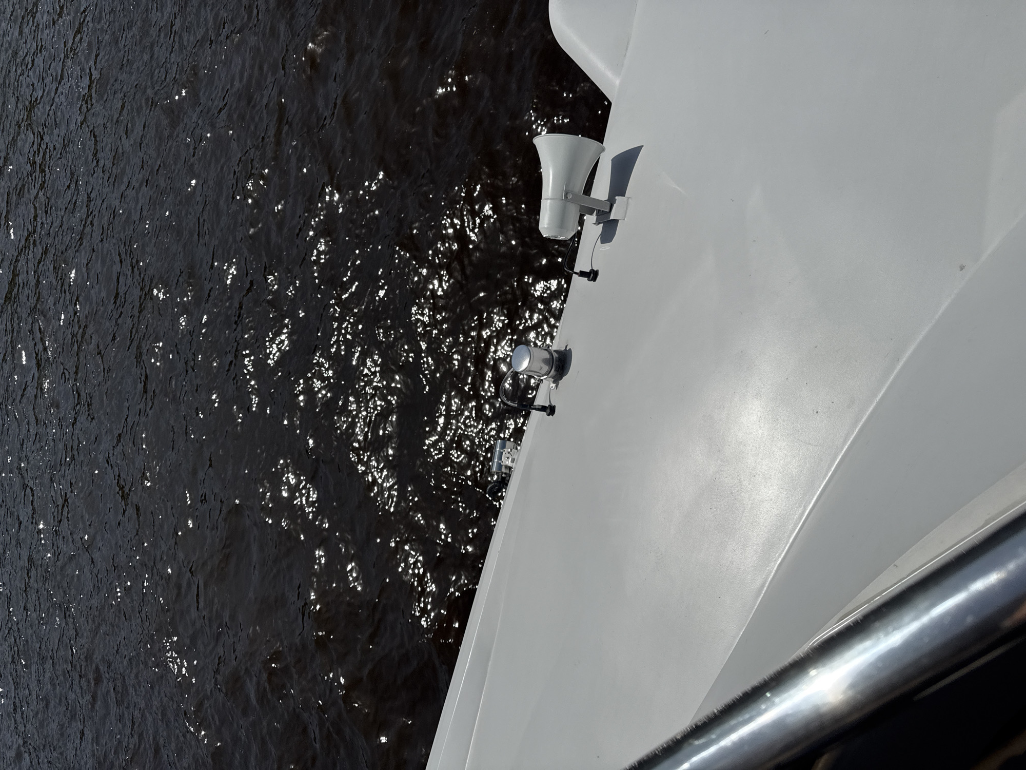 The image depicts a view from a vessel, likely a boat or ship, on the Volga River in Nizhny Novgorod, Russia. The dark, reflective surface of the water occupies a significant portion of the left side of the frame, with scattered light reflections across its surface. The right side of the image is dominated by the white hull of the vessel. Mounted on the hull are two metal fixtures: a white public address speaker and a metallic, cylindrical object. A railing partially obscures the lower right corner of the image. The scene is shot from a high angle, looking down at the river and the vessel's edge.