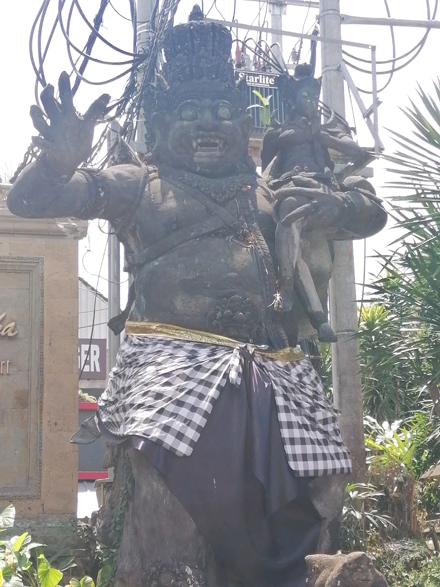 A large stone statue of a demon carrying a smaller figure stands outdoors, possibly in Ubud District, Indonesia, given its Balinese style. The demon has a wide, toothy grin, bulging eyes, and wears a dark crown. It is adorned with a gold and black checkered cloth around its waist, over a dark fabric. The smaller figure, also stone, is held in the demon's left arm, and appears to be a child or a deity. The statue is positioned in front of a stone wall with intricate carvings and signage, partially obscured by the statue and surrounding tropical foliage, including palm fronds and other green plants. Overhead, a complex network of electrical wires and a utility pole are visible against a bright sky. The lighting suggests it is daytime, with bright sunlight casting shadows.