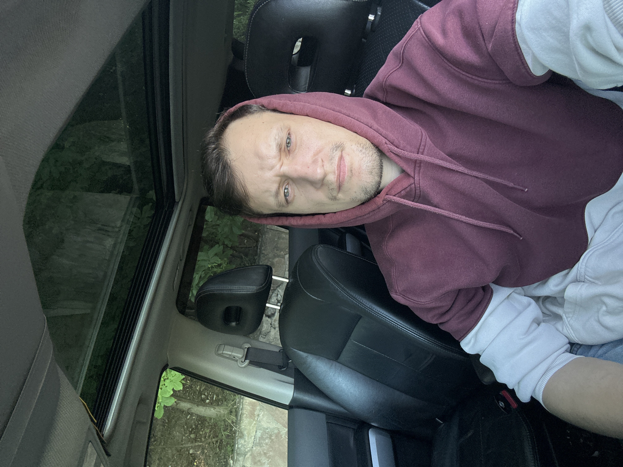 A man is seated in the driver's seat of a car, with a sunroof open above him. He is wearing a maroon hooded sweatshirt over a white long-sleeved shirt. The interior of the car is visible, including black leather seats and seatbelts. Outside the car, trees and foliage are visible, suggesting a natural or wooded environment. The man is looking directly at the camera with a slightly furrowed brow. The scene is illuminated by natural light. There are no visible indications of the specific location within Makiv, Ukraine, beyond it being a place with trees and a car.