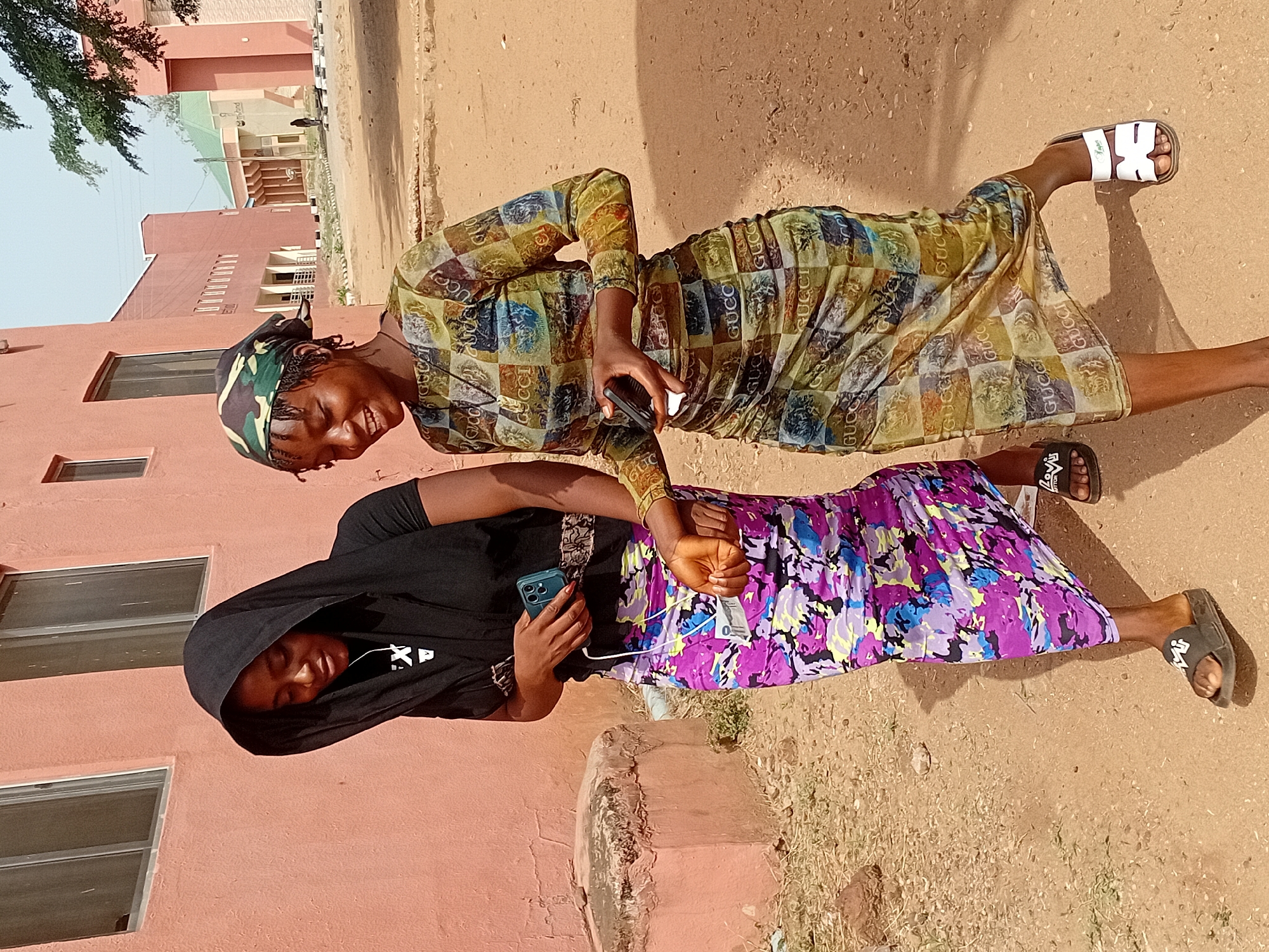 Two young African women are standing on what appears to be a dirt road or sidewalk in Lokoja, Nigeria. One woman is wearing a Gucci print dress, a patterned head covering, and white sandals. She is smiling and holding a cell phone. The other woman is wearing a black hooded sweatshirt, a floral print skirt, and black sandals, and she is also holding a cell phone with earbuds hanging from it. They are standing close to each other, suggesting a friendly interaction. In the background, there are buildings with a pinkish hue and windows, suggesting an urban setting. The ground is predominantly a light brown, earthy color.