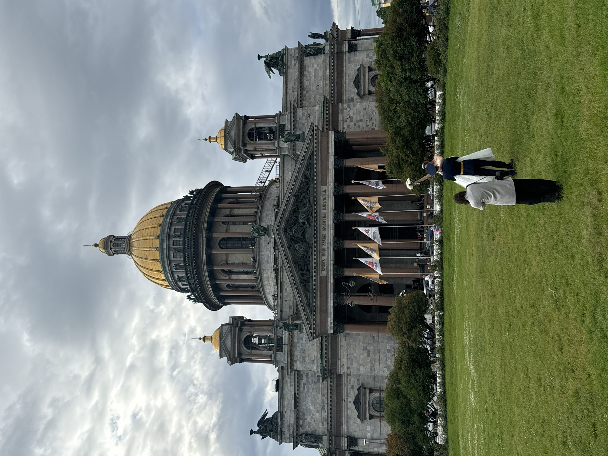 The image depicts a wedding couple posing for a photograph in front of St. Isaac's Cathedral in Saint Petersburg, Russia. The couple is positioned on a grassy area, with the bride in a white dress holding a bouquet and the groom in a dark uniform with gold trim. A photographer, dressed in a gray jacket and black skirt, is capturing the moment. The cathedral, a prominent landmark with a large golden dome, serves as the backdrop. Several flags are displayed in front of the cathedral, and trees line the perimeter of the grassy area. The sky is overcast.