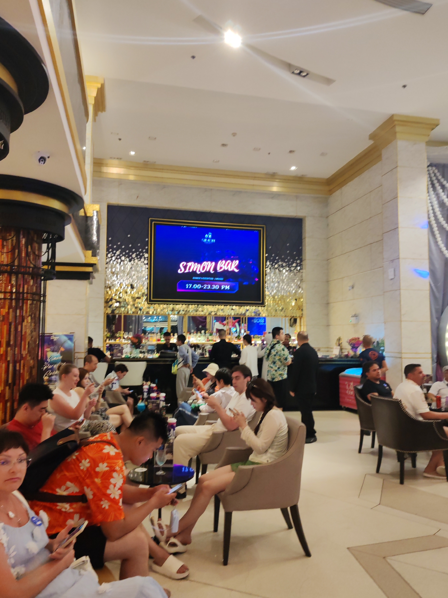 The image shows the interior of Simon Bar in Patong, Thailand. Patrons are seated in chairs, many using mobile devices. A large screen displays the bar's name and operating hours (17:00-23:30). Staff are visible behind the bar. The decor includes ornate columns, marbled surfaces, and ambient lighting.