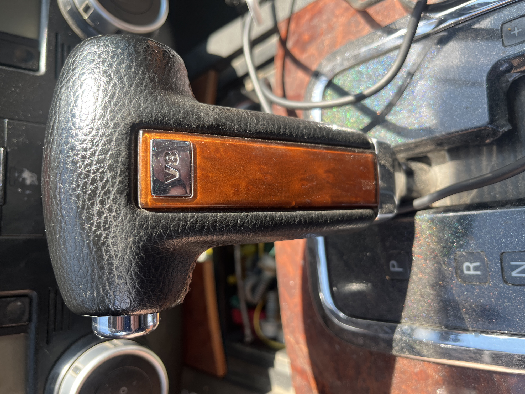 The image shows a close-up of a car's gear shifter and dashboard, which appears to be inside a vehicle located in Aqköl, Kazakhstan. The gear shifter is made of black leather with a wood-grain accent and a "V8" emblem. The dashboard features two circular gauges and a panel with gear indicators "P," "R," and "N." Wires are visible in the background, suggesting the car's interior is somewhat exposed or undergoing maintenance. The lighting indicates it is daytime, with sunlight casting reflections and dust particles on the surfaces.