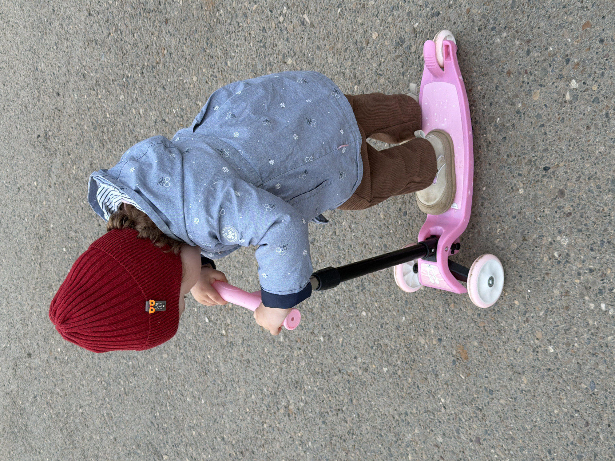 A child wearing a red knit hat, a blue patterned jacket, and brown corduroy pants is standing on a pink scooter with three wheels on a paved surface. The scooter has a black handlebar and pink grips. The child is holding the handlebars and appears to be looking down, but not at the city of Chaikovsky, Russia. The surface appears to be asphalt or concrete. The image is taken from an overhead perspective.