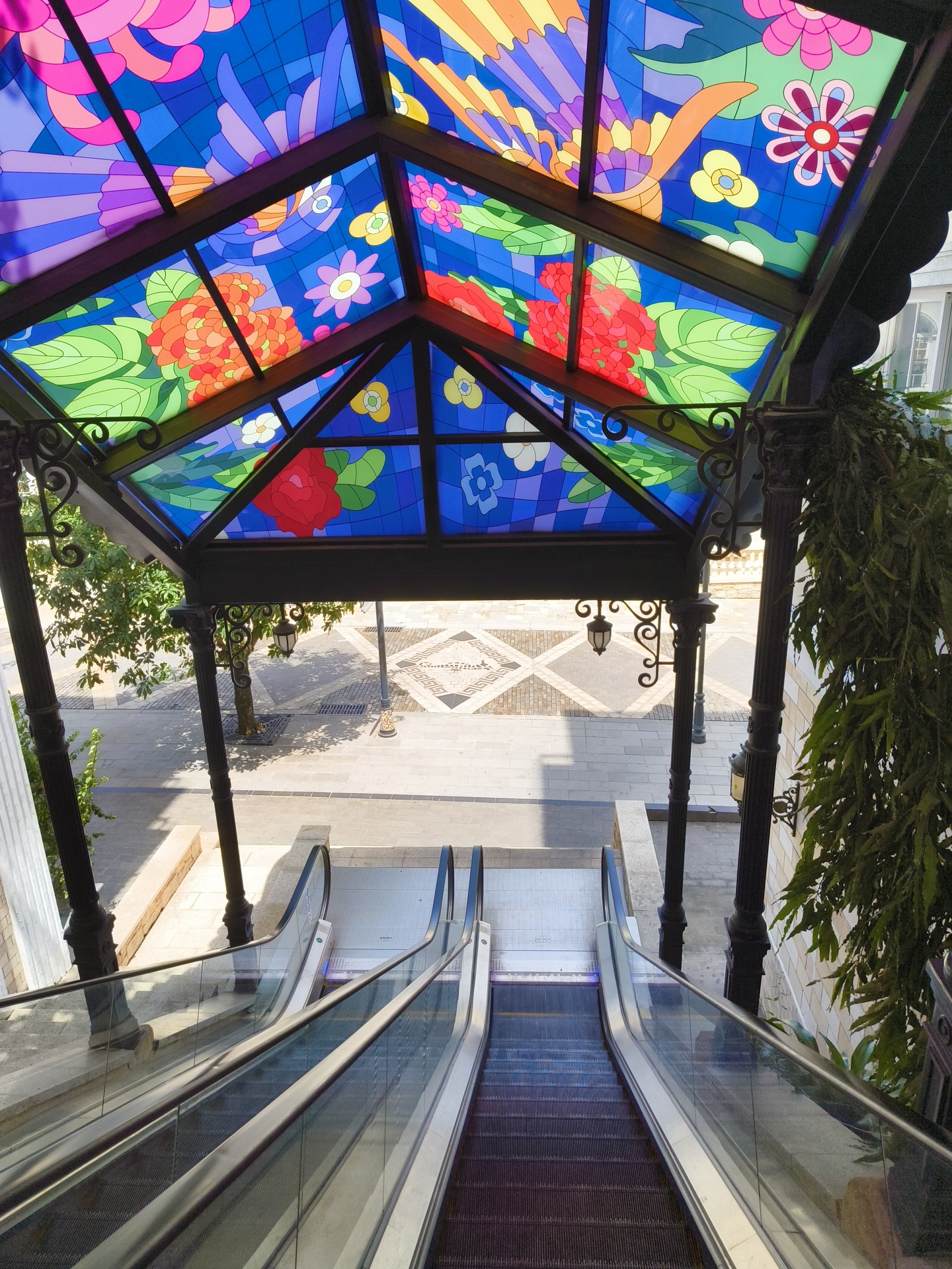 An outdoor escalator in Phu Quoc, Vietnam, leads down from a covered entrance. The entrance features a brightly colored stained-glass ceiling adorned with floral and bird motifs. The escalator is made of metal and glass, and its steps are dark gray. The surrounding environment includes a paved walkway, trees, and architectural elements.