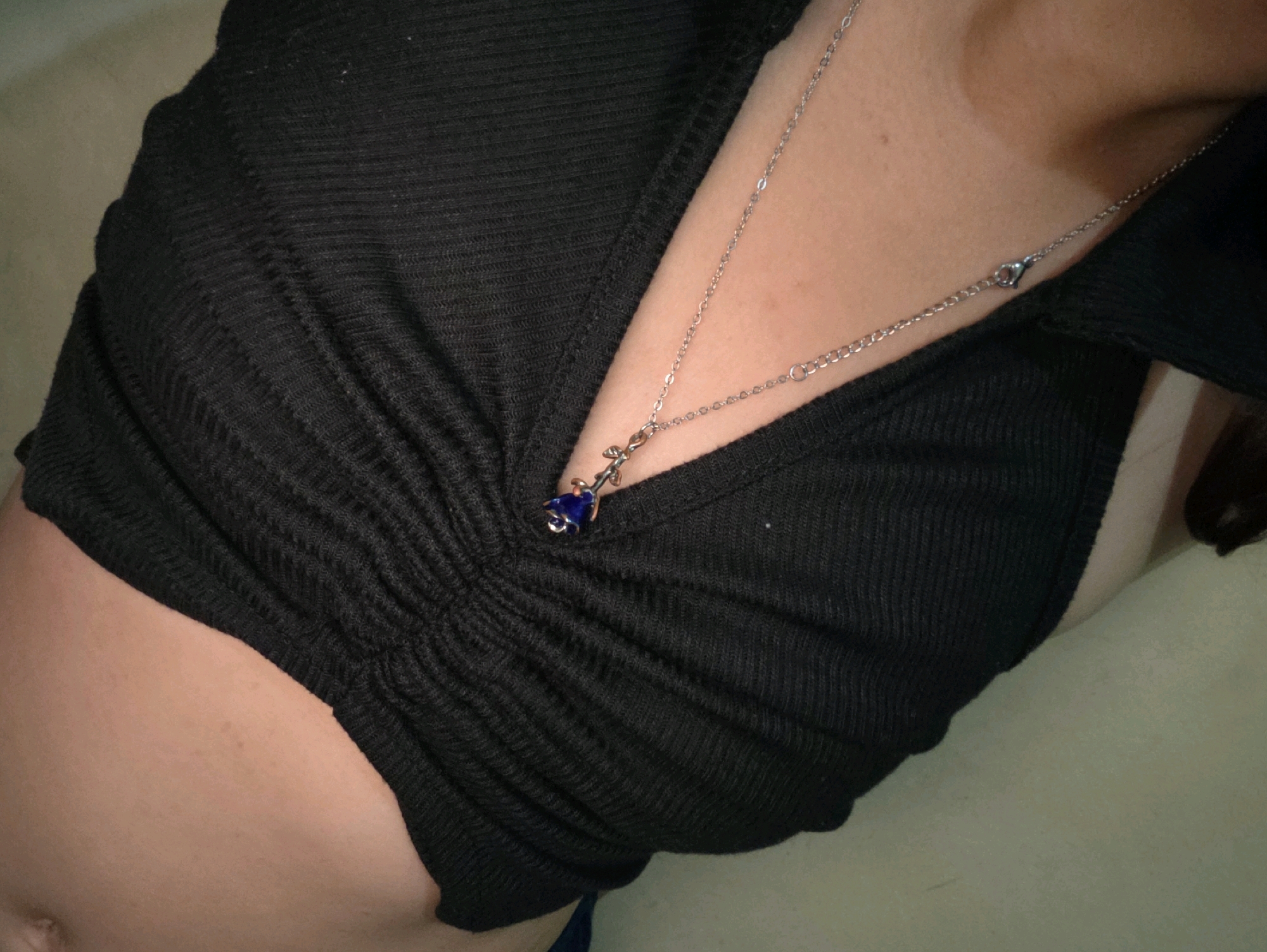 The image is a close-up, overhead shot of a person's torso, wearing a black, ruched top and a delicate silver necklace with a small blue pendant. The background appears to be a textured, light-colored surface. The lighting is diffused, creating soft shadows. There is no discernible connection to the city of Phan Thiet, Vietnam.