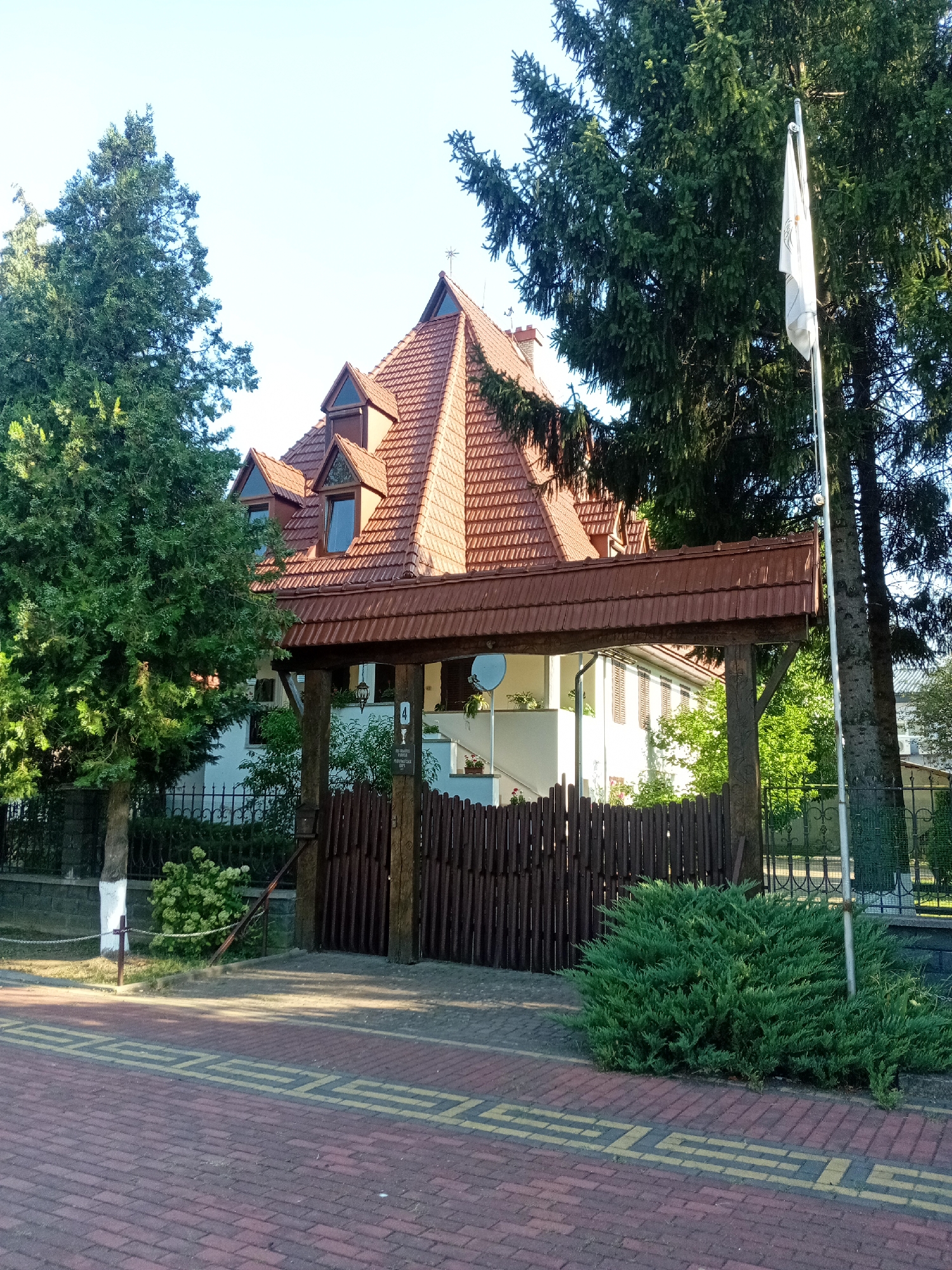 The image depicts the entrance to a building, likely a private residence or institution, in Tiachiv, Ukraine. The architectural style of the building is characterized by a prominent, steeply pitched red-tiled roof with multiple dormer windows. A wooden gate structure with a covered, tiled roof spans the entrance, leading to a fence and the building itself. The fence is made of dark wooden pickets.
In the foreground, a red brick sidewalk with a yellow patterned border runs across the bottom of the frame. To the right of the sidewalk, a well-maintained green shrub is situated. A flagpole with a white flag is visible behind the shrub and to the right of the gate.
The environment appears to be a bright, sunny day. Mature evergreen trees frame the scene on the left and behind the building. The ground beyond the fence is landscaped with greenery and some flowering plants. A white and gray stone wall with a black metal fence separates the property from the sidewalk area. There are no people visible in the image.
