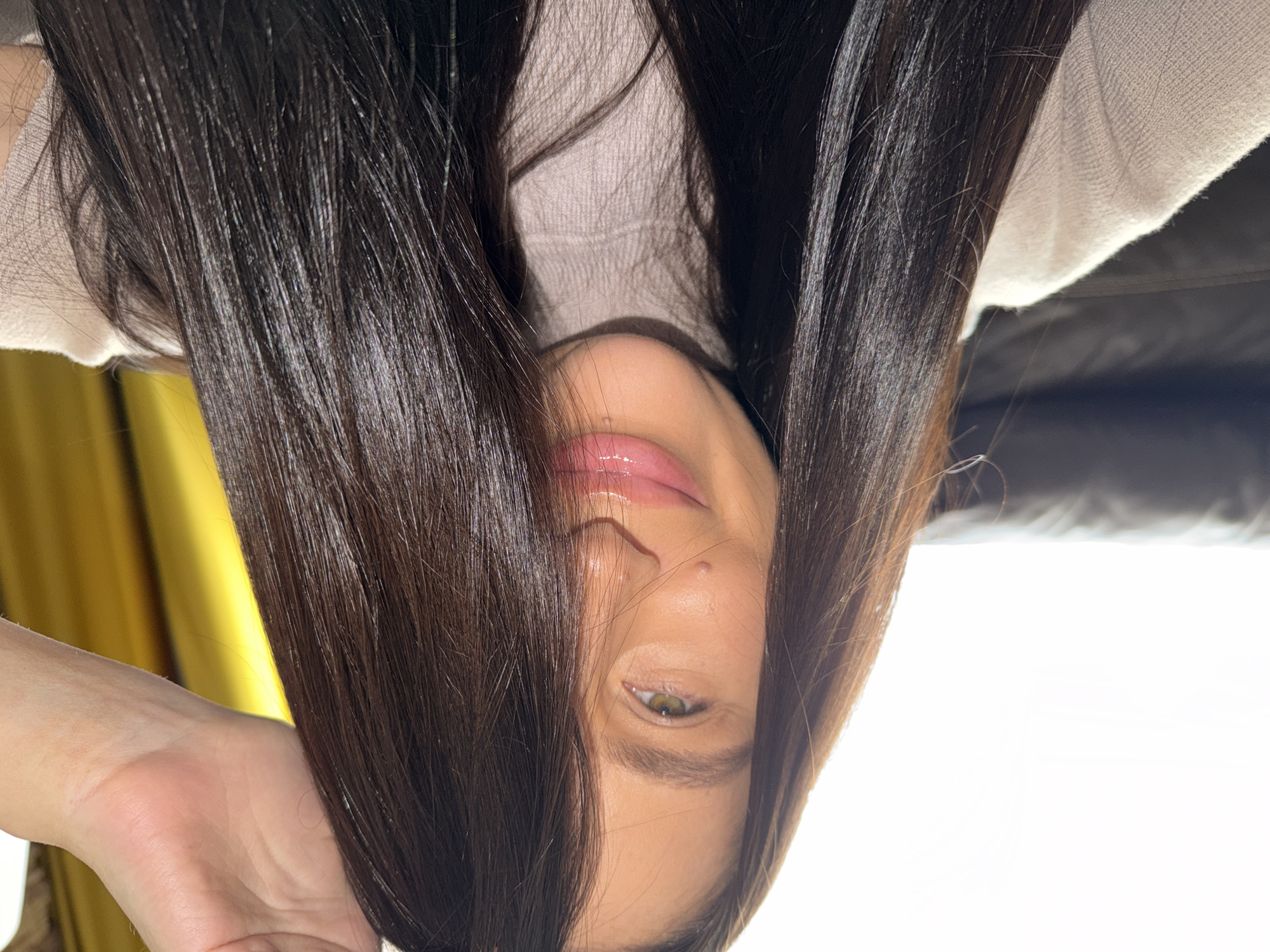The original image is rotated 90 degrees counter-clockwise, presenting the subject upside down. When viewed with correct orientation, the media file presents a close-up portrait of a person, likely female, captured indoors in Khimki, Russia. Her long, dark brown hair, which appears straight and shiny, frames her face as it cascades around her, partially obscuring her features.
Only segments of her face are visible: her glossy, pinkish-red lips are slightly parted, suggesting a relaxed expression. A small mole is noticeable near her nostril. One striking green or hazel eye is visible and looks directly towards the viewer, conveying a serene or thoughtful mood. Her complexion is fair and smooth.
She is dressed in a light-colored, possibly beige or cream, knitted top or sweater. A darker, possibly upholstered surface is visible behind her upper right shoulder. The background includes a golden or mustard yellow curtain on the left, suggesting a nearby window. The right side of the frame is dominated by a bright white area, indicative of strong natural light, likely from another window. The overall lighting suggests the photo was taken during the daytime. There is no visible text in the image.