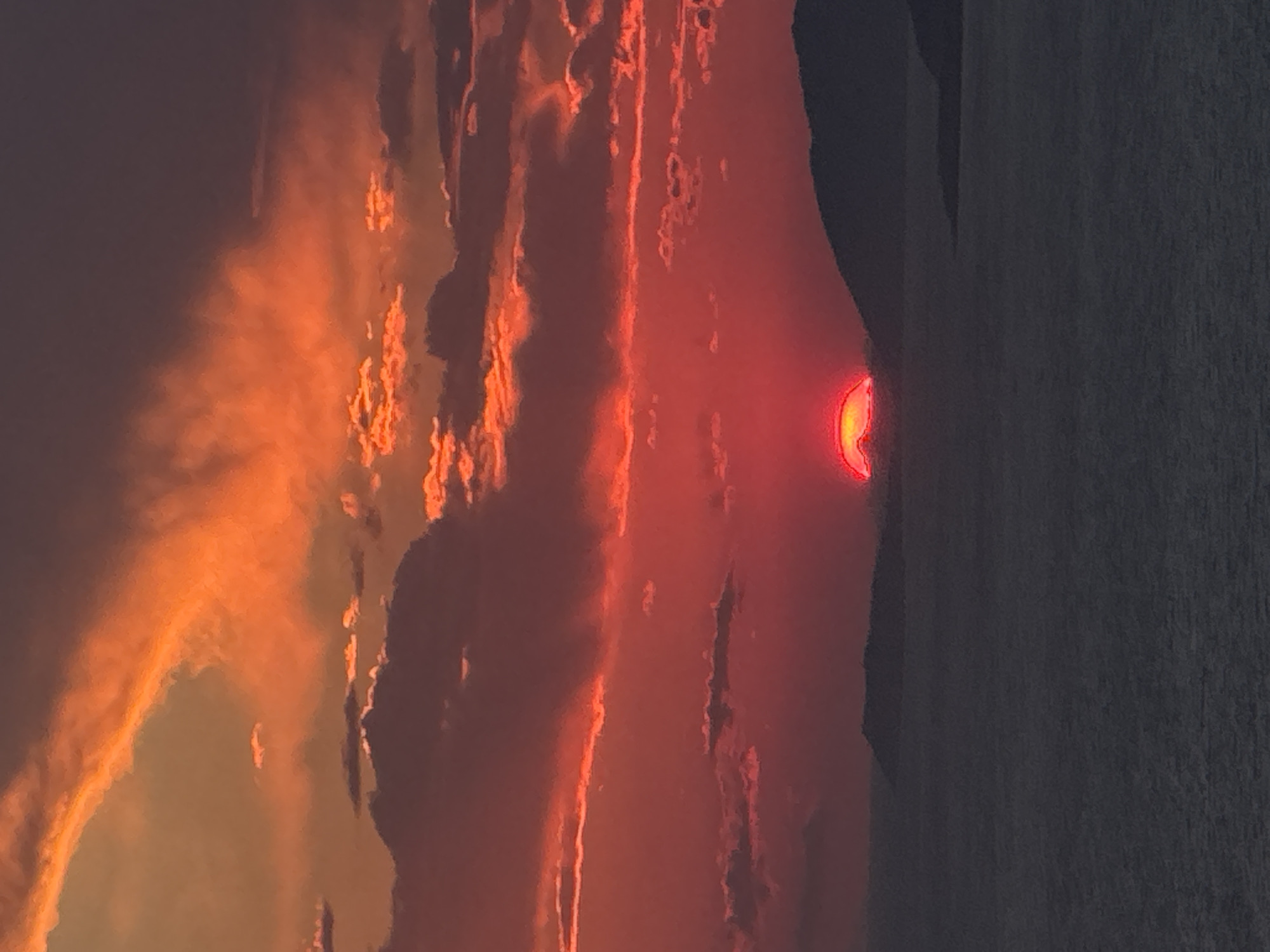 The image depicts a dramatic sunset over the water, with the sun partially obscured by hills and islands. The sky is ablaze with vibrant shades of red and orange, with dramatic clouds casting shadows across the scene. The water in the foreground is dark and appears calm, reflecting the colors of the sky. Silhouetted against the vibrant sky are the dark shapes of hills and islands. The image is a landscape photograph, likely taken from an elevated viewpoint looking towards the horizon. The location is identified as the city of Zagora, Montenegro. There are no people or specific objects visible in the scene beyond the natural elements of the landscape and the sunset.
