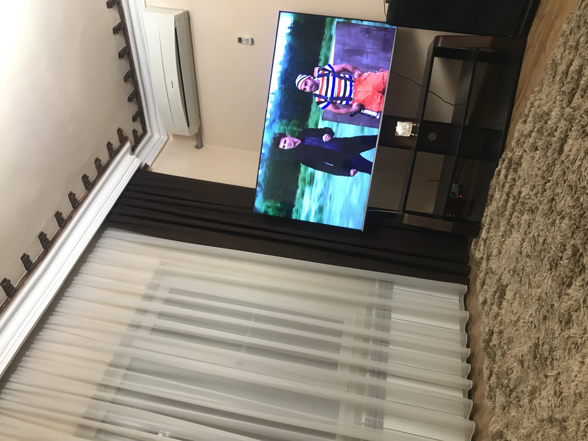 Here is a detailed description of the image:
The image is an interior shot of a room, likely a living room, in Yerevan, Armenia. On the wall is a large flat-screen TV showing a comedy television show. The TV sits on a dark brown entertainment stand. To the left of the TV are curtains that have brown blackout curtains on the top and white sheer curtains on the bottom. Above the TV there is an air conditioner. There is a thick, shaggy rug that covers the floor. There is a decorative molding on the ceiling.