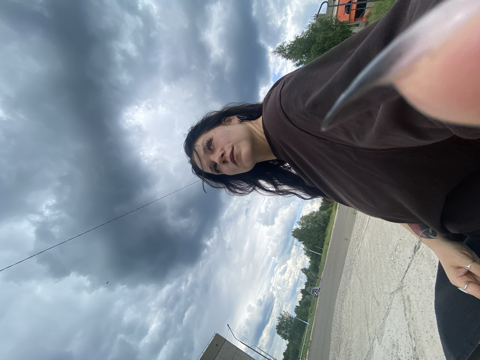 The image is a low-angle, close-up, selfie-style photo taken outdoors in what appears to be a rural or semi-rural setting in Марковский, Russia. The main subject is a young woman with long, dark hair, wearing a brown long-sleeved shirt. She is looking towards the camera with a neutral expression. Her face is partially obscured by her hair and the angle of the shot. Notable details visible include a circular earring in her left ear, a nose ring, and a tattoo on her left forearm, which appears to be black and red. She is wearing two silver rings on her right hand.
The background consists of a dramatic, cloudy sky, suggesting overcast or possibly stormy weather. There are trees and a road visible in the distance, with some man-made structures, possibly part of an industrial or residential area, also present. The ground in the foreground is a cracked concrete surface. A thin, dark line, likely a power line or wire, stretches diagonally across the upper portion of the image. The overall impression is of a casual, candid moment captured outdoors, with the weather contributing to a somewhat moody atmosphere. The lighting suggests it is daytime, but the heavy cloud cover makes it difficult to determine the exact time. No text is visible in the image.