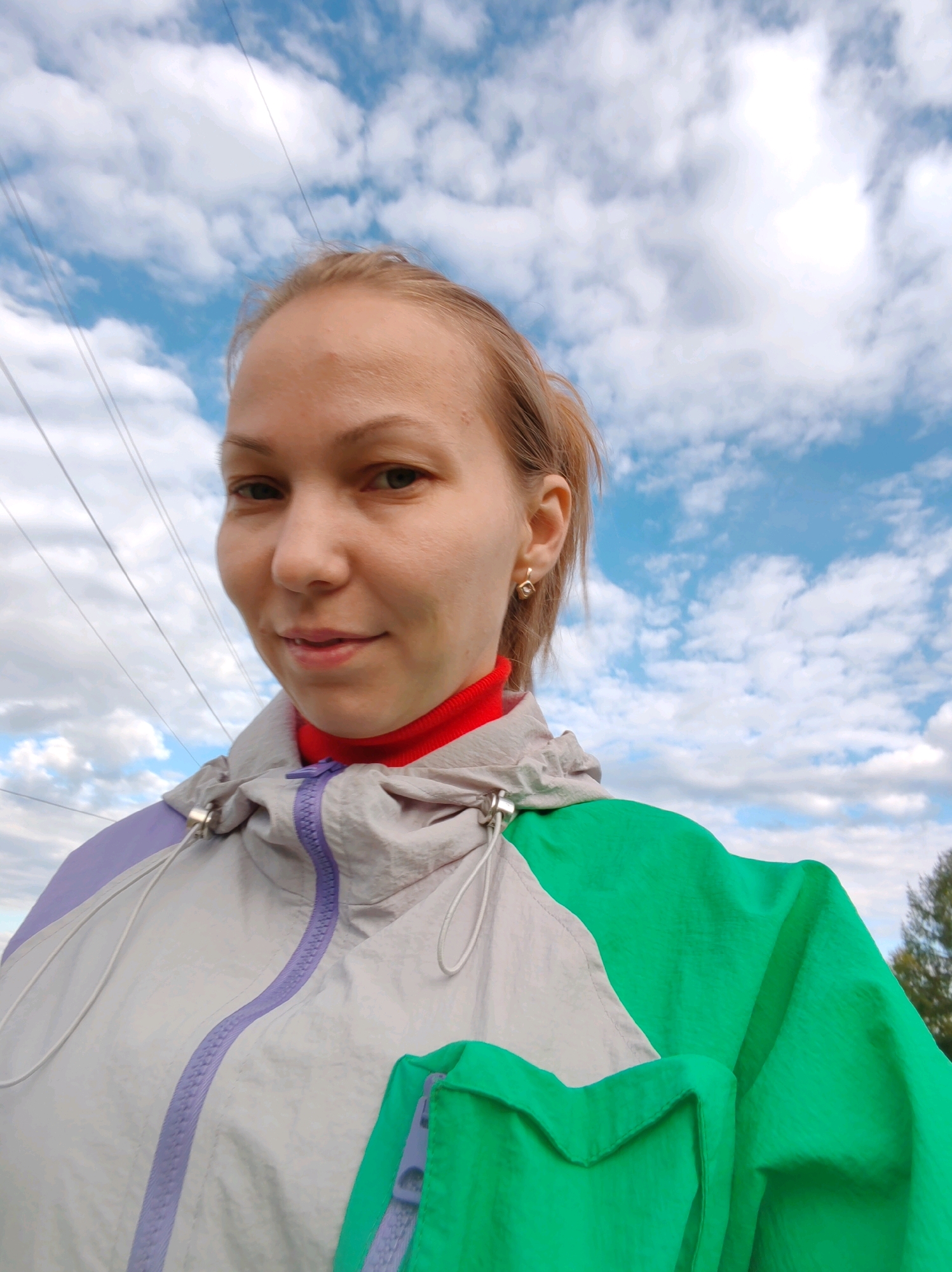 A medium shot photo shows a woman with blonde hair wearing a red turtleneck and a light gray and purple jacket with green accents. She is outdoors against a bright blue sky with white clouds. Several thin, dark wires are visible crisscrossing the sky behind her. She is looking towards the camera and smiling slightly. The photo appears to be a selfie, taken from a low angle. The environment suggests a rural or semi-rural setting, possibly in the city of Chuplem, Russia.