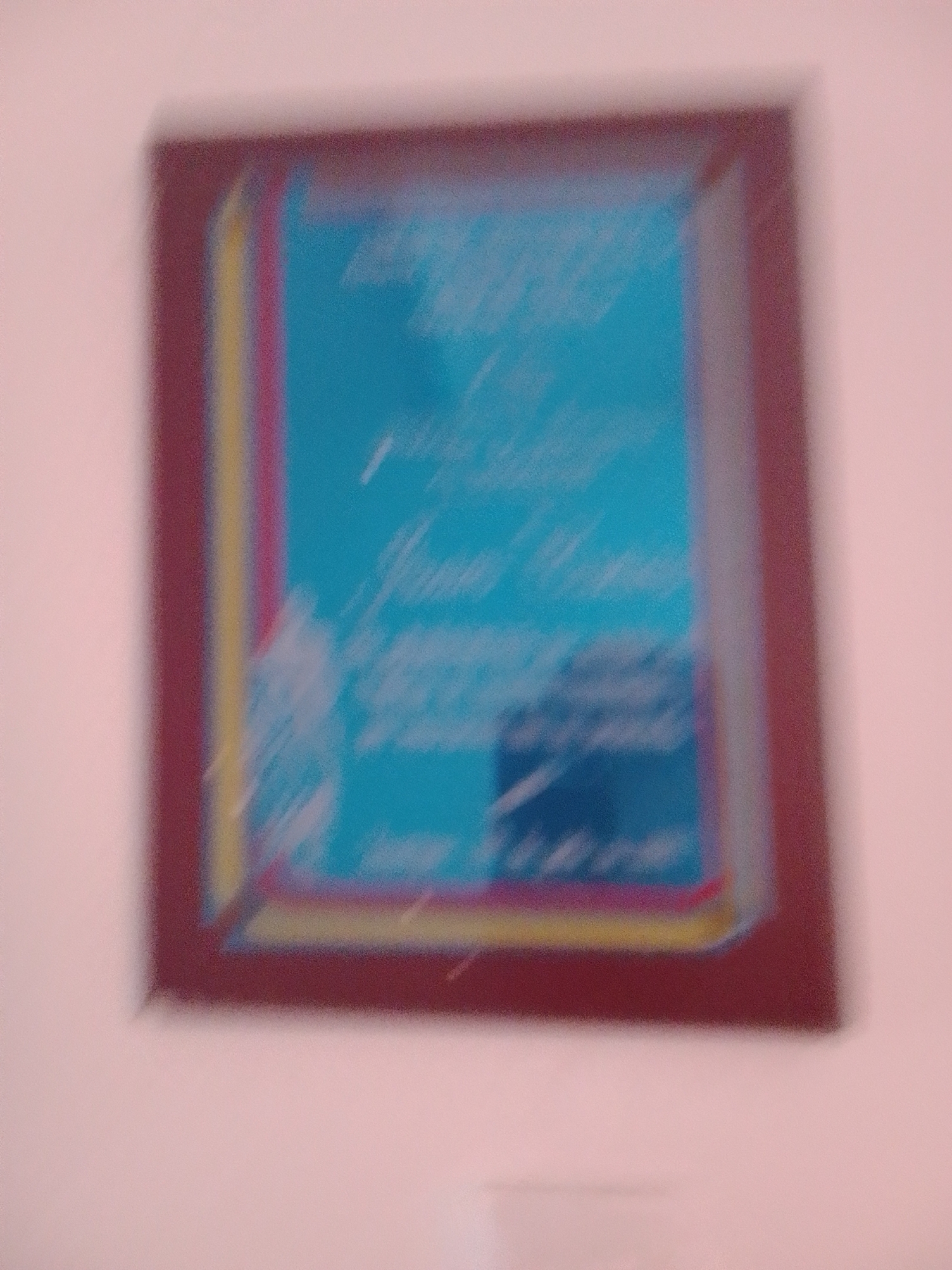 The image shows a framed certificate or award displayed on a wall. The frame has multiple layers of color, with a deep red outer border, followed by a yellow and then a pink inner border, surrounding a blue matting. Inside the matting, there is a light blue insert with white text and some decorative elements. The text is too blurry to be read clearly, but it appears to be a formal document, possibly an award or certificate of recognition. The lighting in the image is soft, suggesting it was taken indoors during the day, perhaps in a home or office setting. The overall impression is of a proud display of achievement.