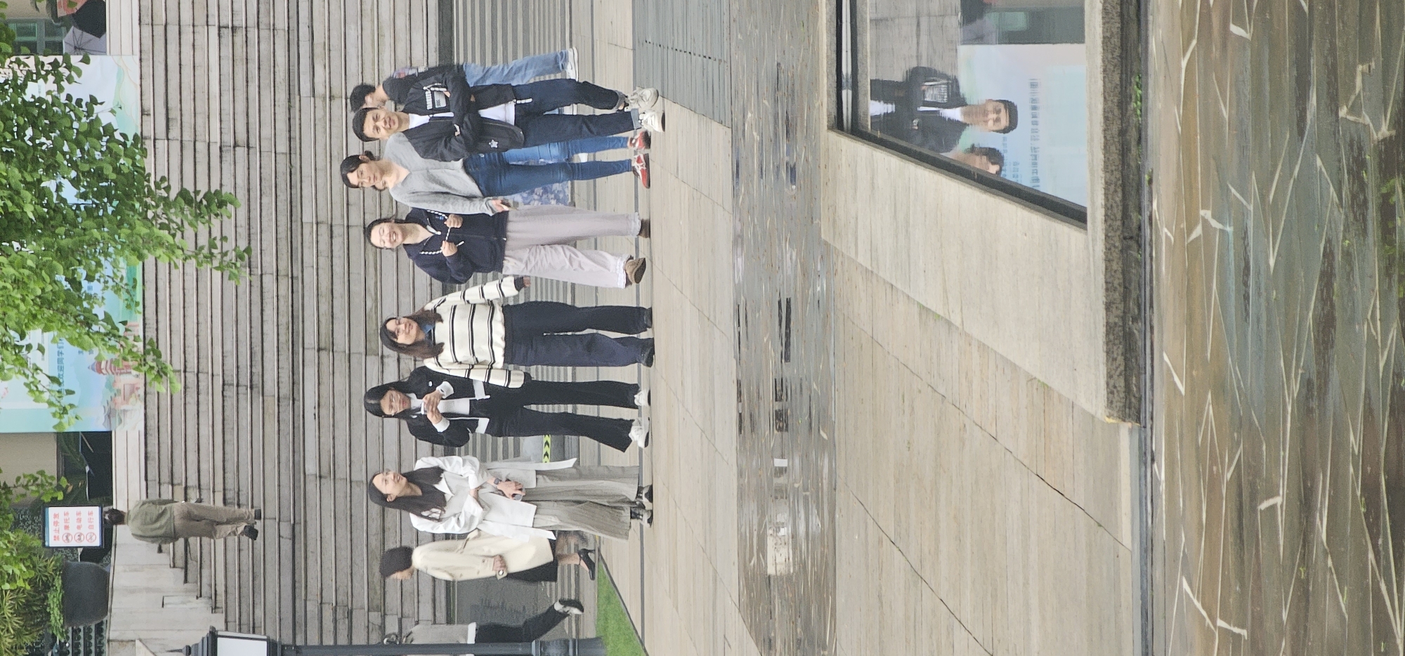 A group of eight people are standing on a staircase in an urban area. The people appear to be young adults, and they are arranged roughly in a line. They are wearing a variety of clothing styles.
The staircase is made of light-colored stone or concrete. A building with a reflective window is to the right of the people. A tree is to the left of the staircase. A man is walking up the stairs. The location is Yuzhong District, China.