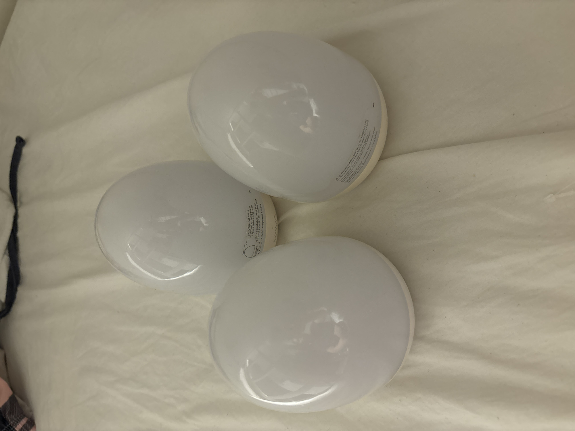 Three white, egg-shaped lamps are arranged on a white, creased fabric surface, likely a bedsheet. The lamps are illuminated and appear to be made of a translucent material. Some text is visible on the base of the lamps, indicating product information and manufacturing details, but the text is not fully legible. The lighting suggests an indoor environment. The image does not contain any information to indicate its location in Dubai, United Arab Emirates.