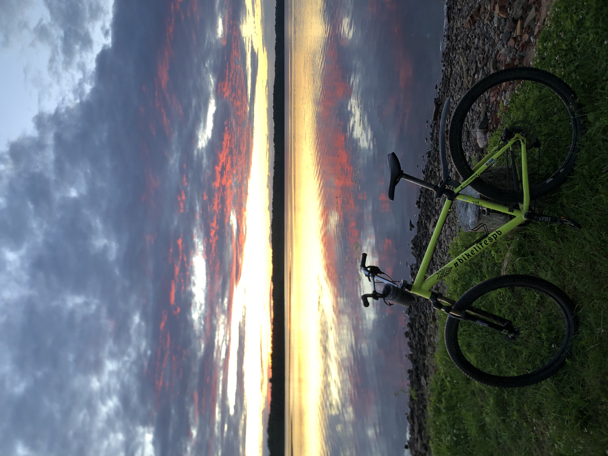 A neon green bicycle rests on a grassy bank next to a body of water at sunset. The sky is filled with dramatic clouds, tinged with pink and orange from the setting sun. The water reflects the colorful sky and the distant tree line. The bicycle is facing the water, with its front wheel on the grass and its rear wheel on a rocky shore. The scene appears peaceful and serene, with no people visible. The location is the city of Svirstroy, Russia.
