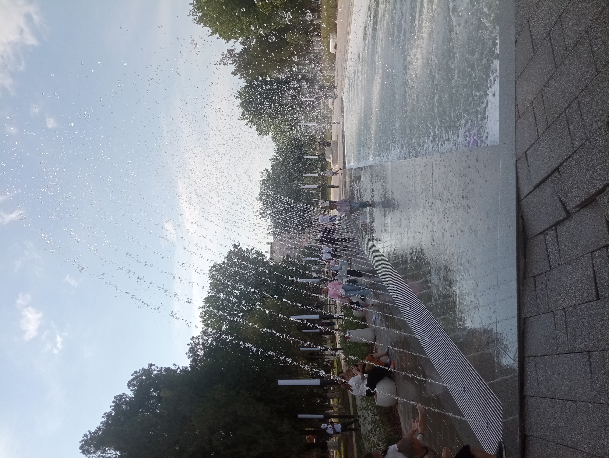 The image depicts a modern public fountain in Shymkent, Kazakhstan. The fountain features a large, flat water surface with numerous thin jets of water spraying upwards, creating a dynamic and refreshing display. The water cascades down in sheets and streams, catching the light and creating a shimmering effect.
Several people are present, enjoying the fountain on a sunny day. Some are standing near the edge of the water, while others are seated on benches or the surrounding pavement. Children appear to be interacting with the water, with one person in the foreground reaching out a hand towards a spray. The overall atmosphere is one of relaxation and enjoyment, with people taking advantage of the cool water to escape the warmth of the day.
The setting includes lush green trees framing the fountain area, suggesting a park or public square. The ground is paved with textured, dark gray tiles, and there are white cylindrical objects that might be decorative lights or part of the fountain's infrastructure. The sky is a bright blue with scattered white clouds, indicating fair weather. The time of day appears to be daytime, likely afternoon, given the brightness of the light and the presence of people enjoying outdoor activities.
The image is captured from a low angle, looking up towards the sky and the fountain's spray. The water jets are a prominent element, creating a curtain-like effect across the frame. The reflection of the water and the surrounding environment is visible on the polished surface of the fountain's basin. No visible text is present in the image.