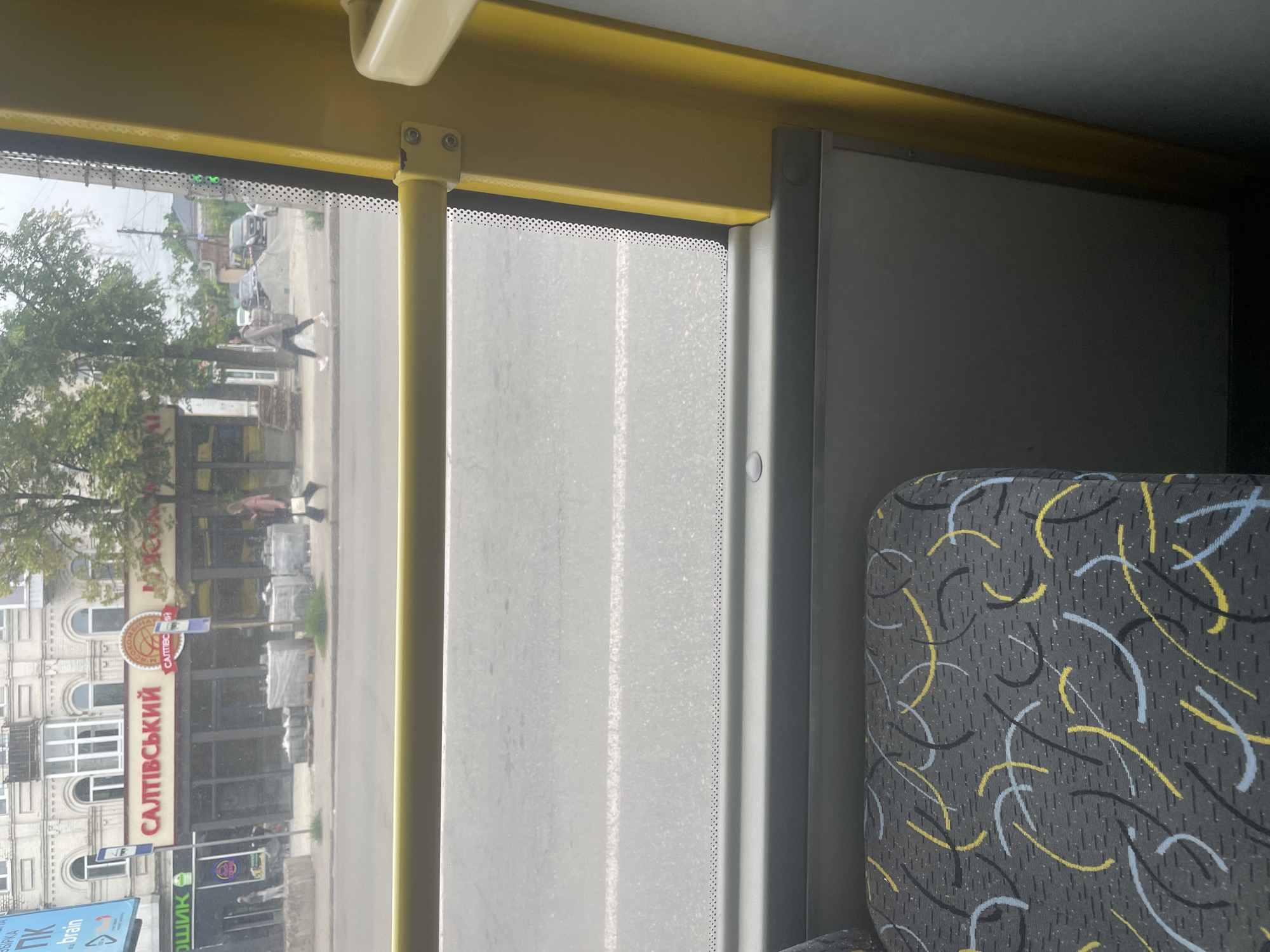 The image, taken from inside a yellow bus, shows a street scene in an urban environment. Through the window, we see a building with signage that reads "САЛТІВСЬКИЙ" in large red letters, and "ЕКОНОМ" in a circular emblem. Next to it, another sign appears to be for a grocery store. People are visible walking on the sidewalk, some carrying bags. Construction materials, such as stacked bricks wrapped in plastic, are present on the sidewalk. A road with a white line marking runs in front of the building. The interior of the bus is partially visible, showing a seat with a patterned fabric and a yellow metal support pole. The overall lighting suggests it is daytime, with overcast skies.