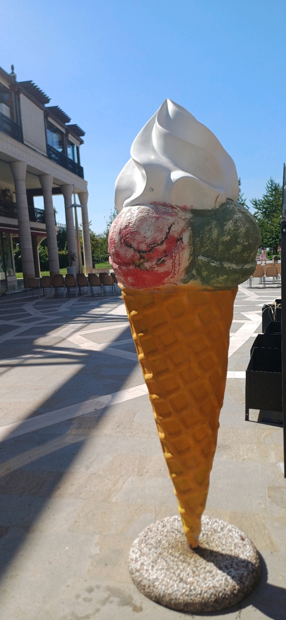 A colossal, weathered sculpture of a three-scoop ice cream cone stands on a circular pebble base in an outdoor plaza. The sculpture features a white, swirled top scoop, a red scoop with dark crackling details, and a green scoop. The cone itself is a bright yellow-orange, textured to resemble a waffle cone. The plaza has a patterned paved ground with geometric shapes and is casting strong shadows from a bright, clear blue sky. In the background, a multi-story building with columns and balconies is visible, along with rows of chairs and trees. This scene is located in the city of Abano Terme, Italy.