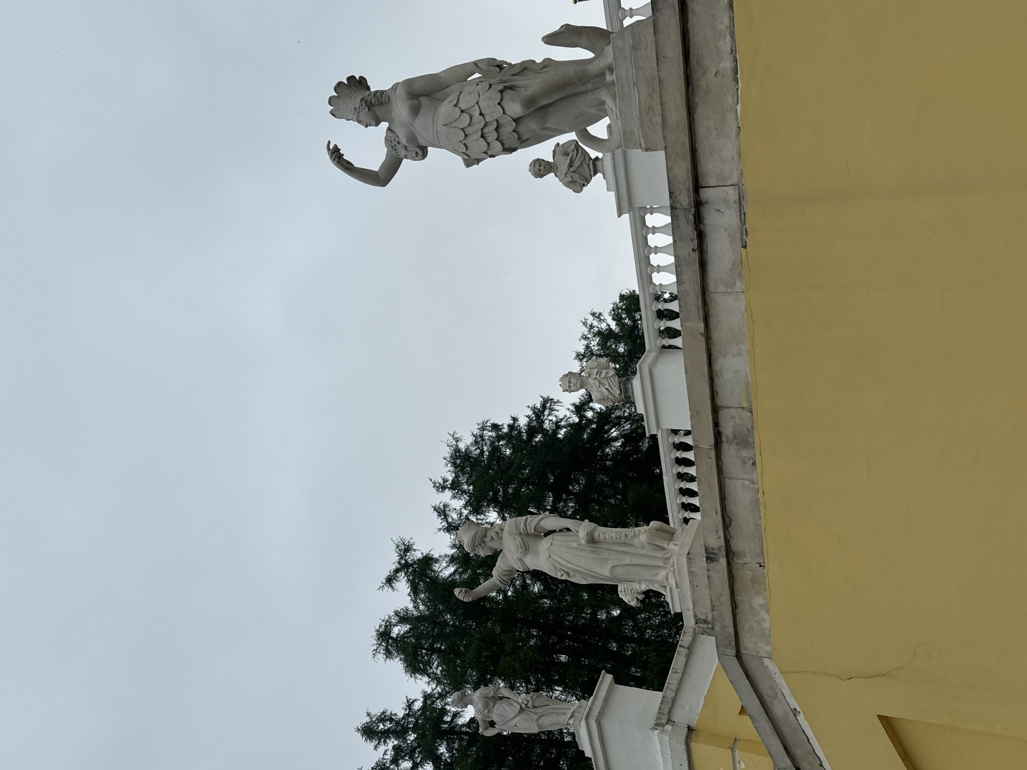 The image shows a low-angle view of a building exterior in Arkhangel'skoe, Russia. The building is painted yellow and features a white balustrade adorned with classical-style statues. Three statues are prominently visible, with a tree appearing in the background behind the lower two statues. The sky is overcast. The statues are pale in color and depict human figures in classical garb.