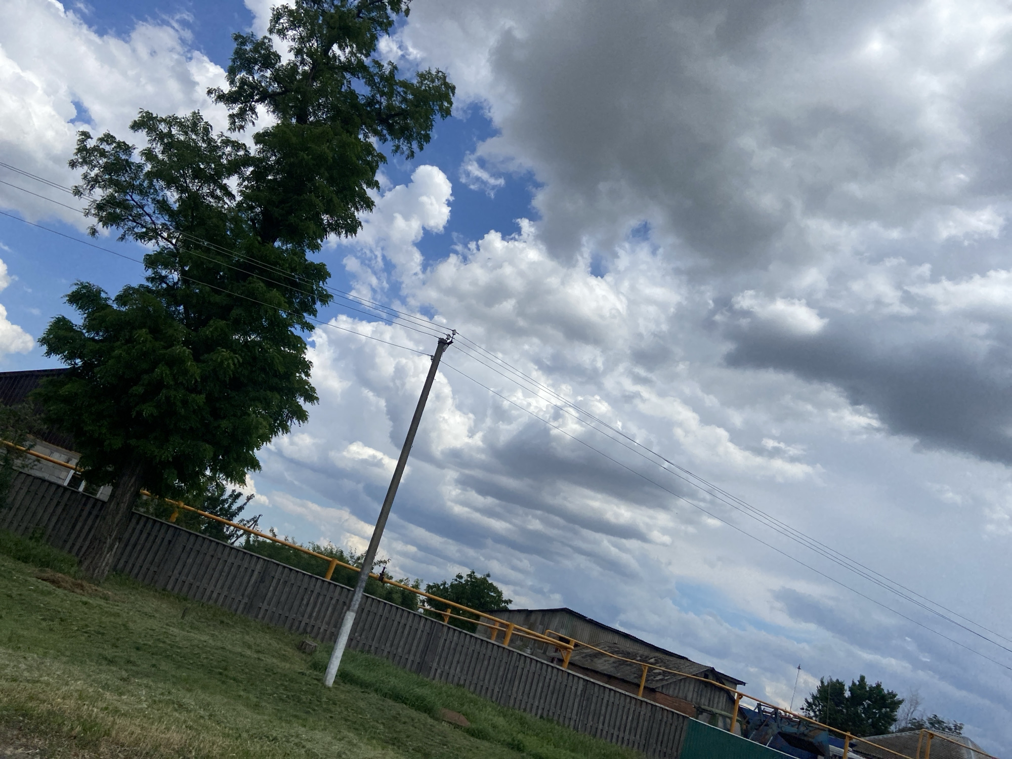 The image depicts a cloudy sky with some patches of blue, above a scene that appears to be in the Tagarogsky district of Russia. In the foreground, a sloped grassy area leads up to a wooden fence topped with a yellow railing. Behind the fence, there are buildings with dark roofs and some trees. A utility pole with wires runs diagonally across the frame. The overall impression is one of a somewhat overcast day in a semi-rural or industrial area.
